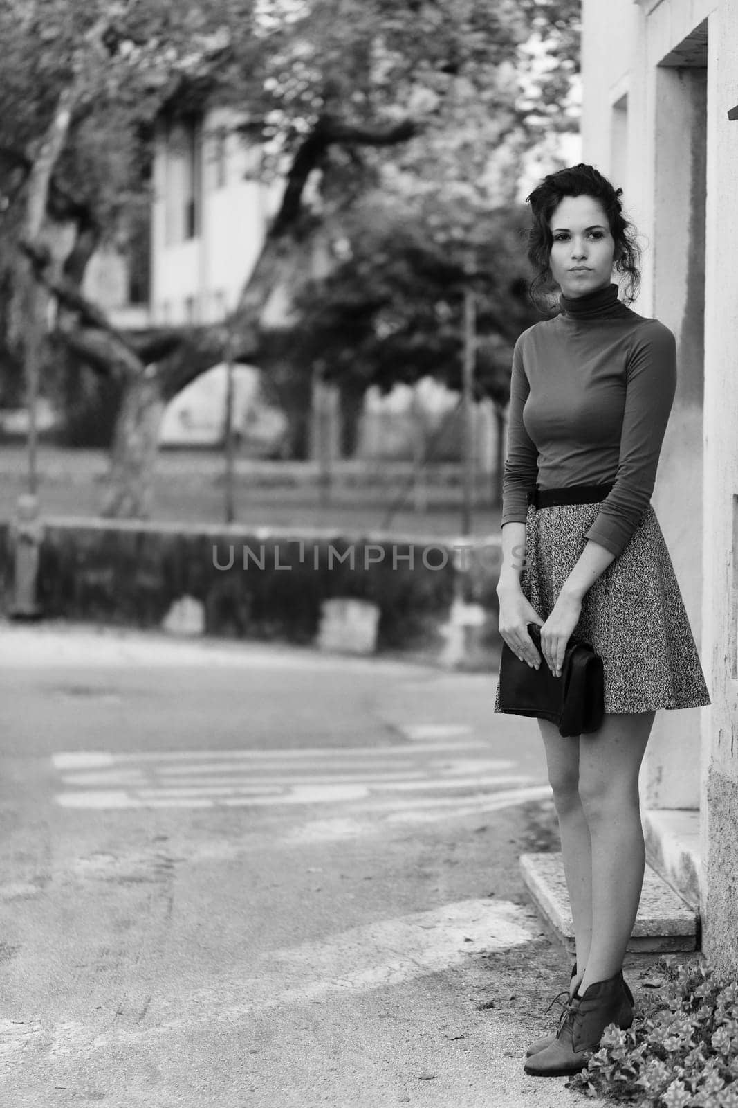 young woman stands near a building, appearing to wait for someone. She wears a brown turtleneck and grey skirt, holding a clutch. The black and white photo features a blurred background of trees