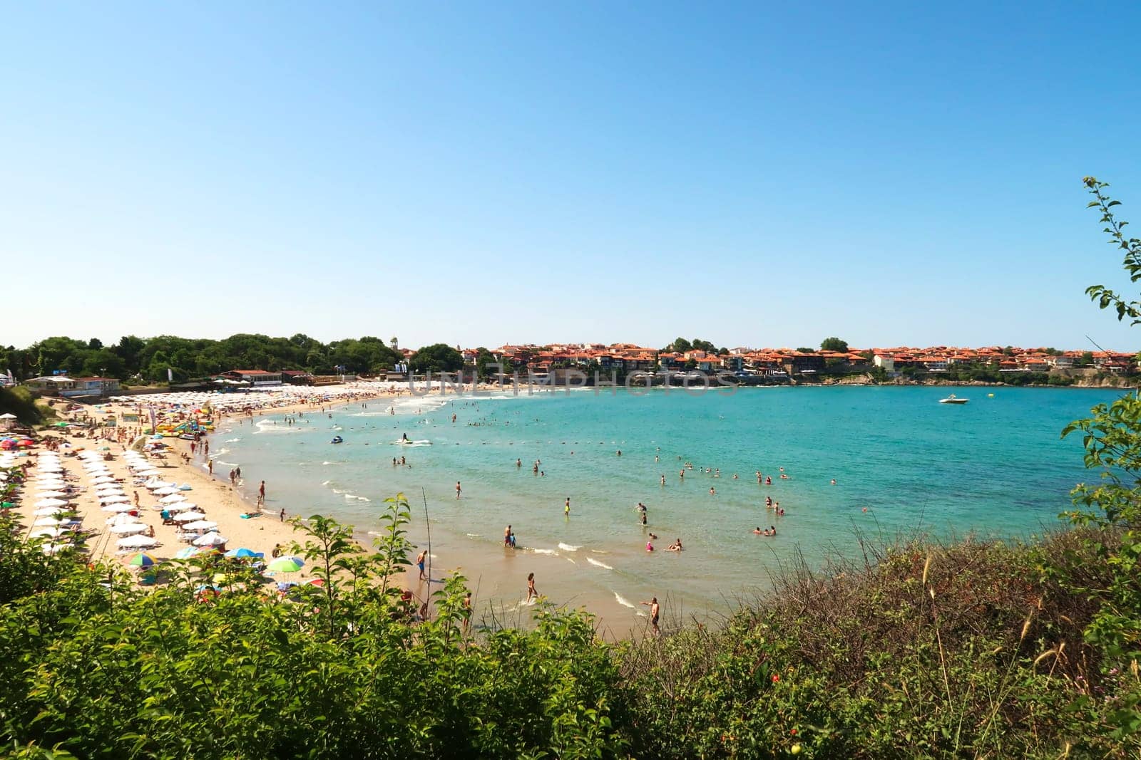 View onto the Sozopol Central Beach on a busy day, white beach umbrellas and sun chairs on the beach, the intense turquoise blue sea and the old town of Sozopol, Sosopol in the background, Bulgaria, 5th of July 2022