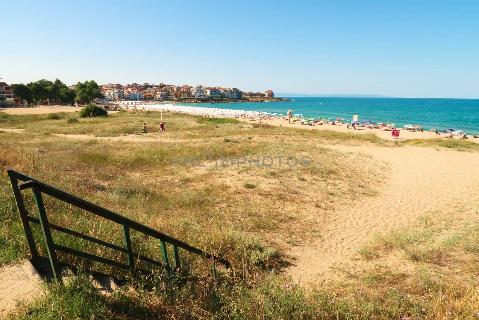 Stairs leading down to Harmanite, Kharmanite on a busy day, many white beach umbrellas and sun chairs on the beach, the intense turquoise blue sea and the town of Sozopol, Sosopol in the background, Bulgaria, 5th of July 2022