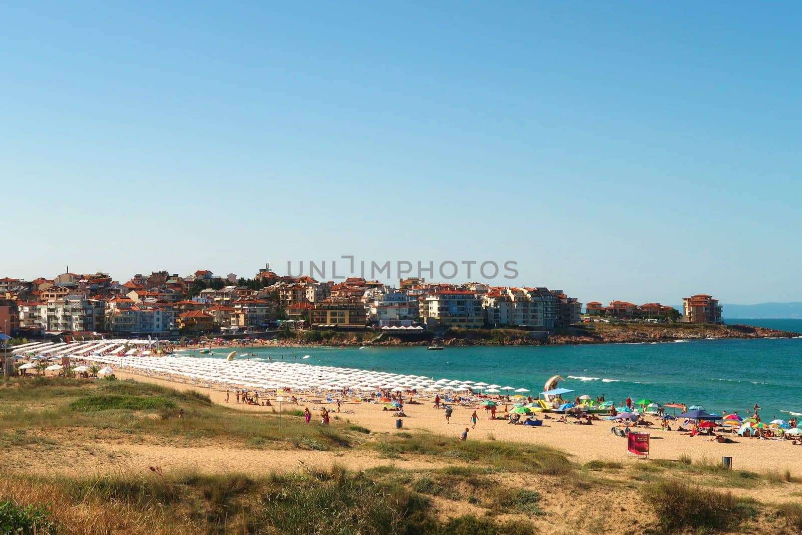 View onto Harmanite, Kharmanite on a busy day, many white beach umbrellas and sun chairs on the beach, the intense turquoise blue sea and the town of Sozopol, Sosopol in the background, Bulgaria, 5th of July 2022