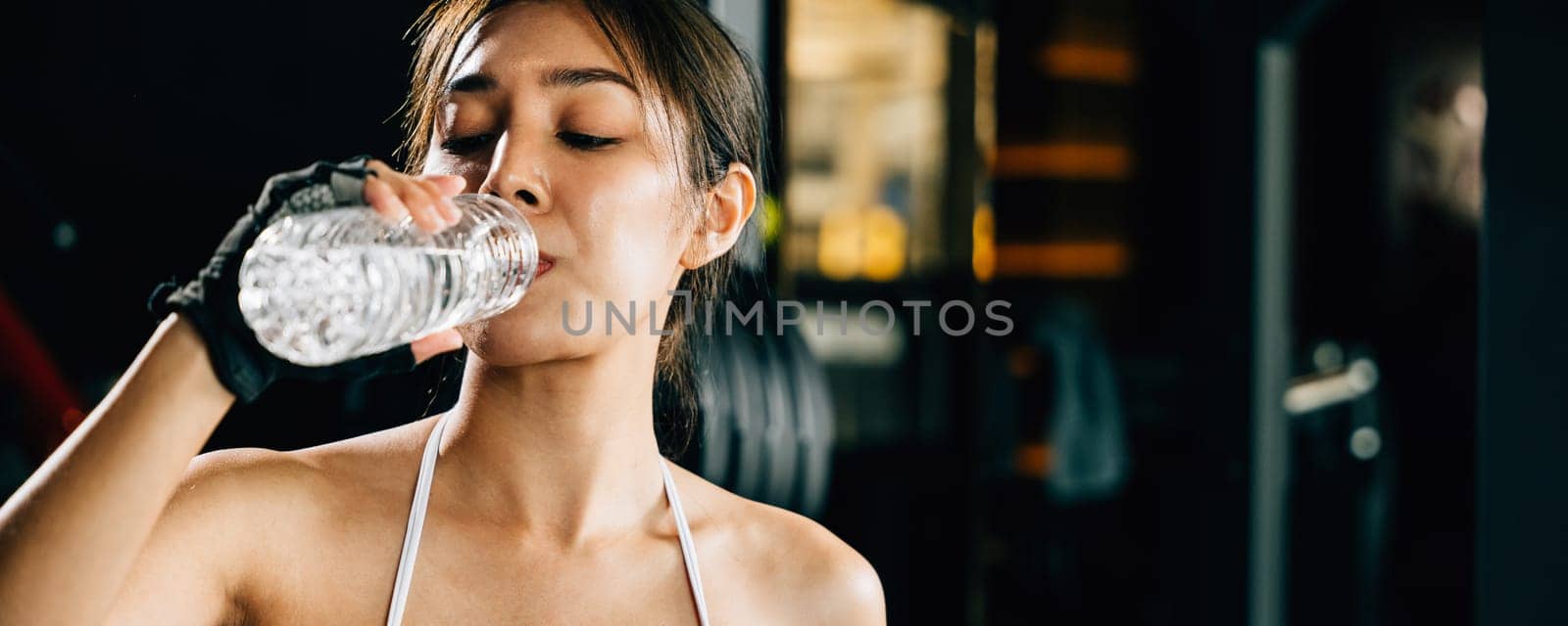 Asian woman guy hydrated by drinking water sports from a bottle in gym after cardio work out, female tired with liquid bottle drink after training at fitness gym, workout healthy lifestyle concept