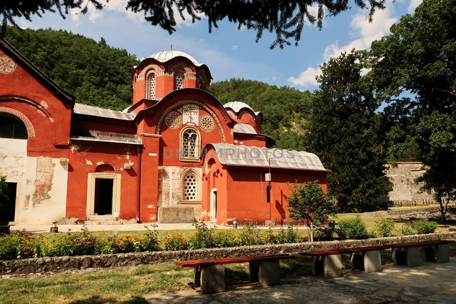 View onto the elaborate front facade and roof, domes, cupolas of the bright red Church Complex inside the courtyard, garden of the Patriarchate of Pec Monastery with a bench in the foreground, outside of Peja, Kosovo 2022