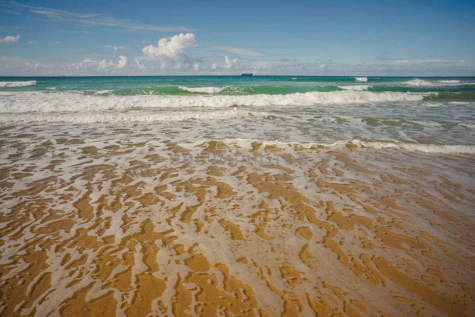 Ocean view with ship on horizon from Kawana Beach, Queensland, with surf, waves, sand and blue cloudy sky in winter.