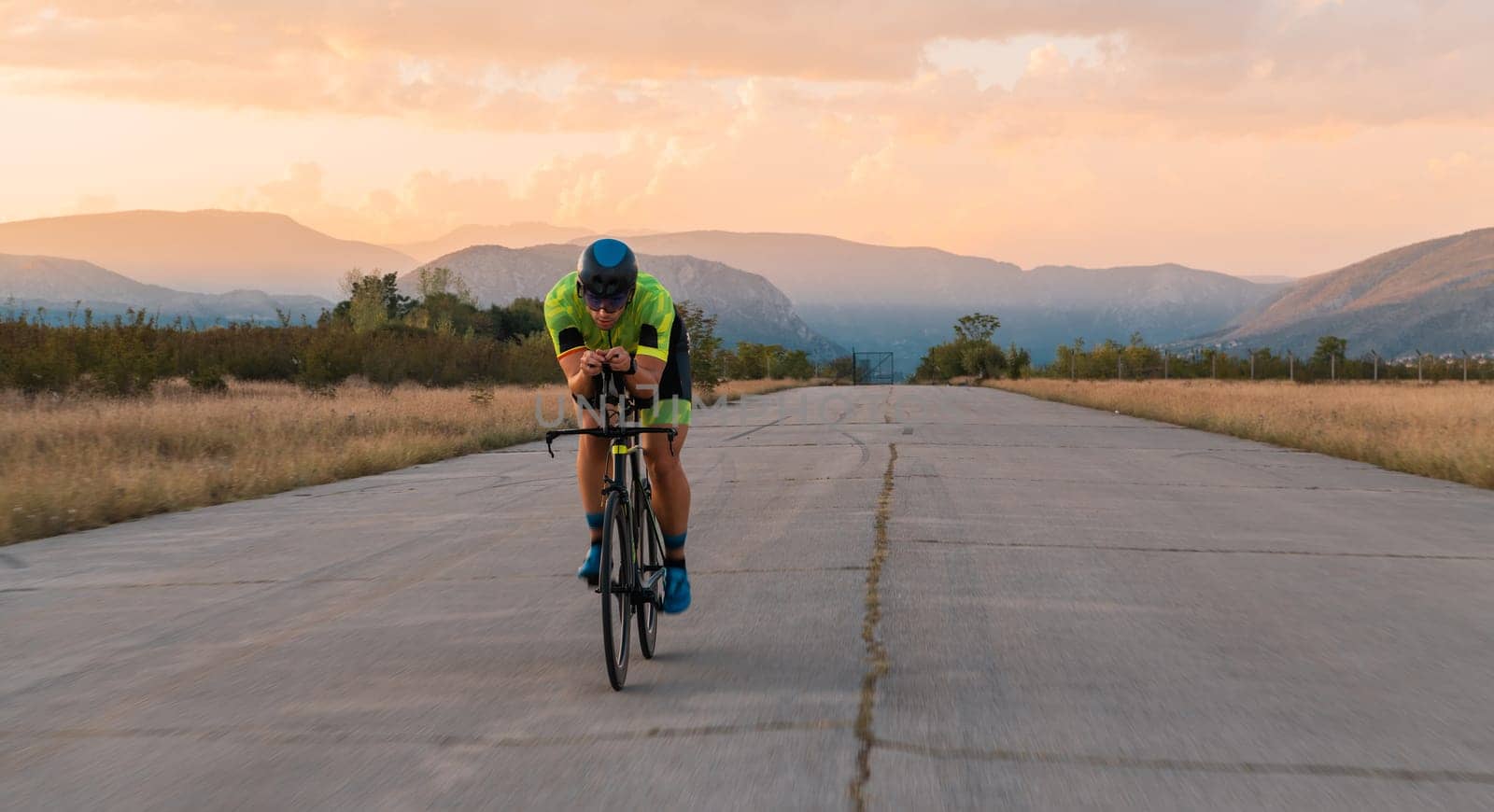 Triathlete riding his bicycle during sunset, preparing for a marathon. The warm colors of the sky provide a beautiful backdrop for his determined and focused effort