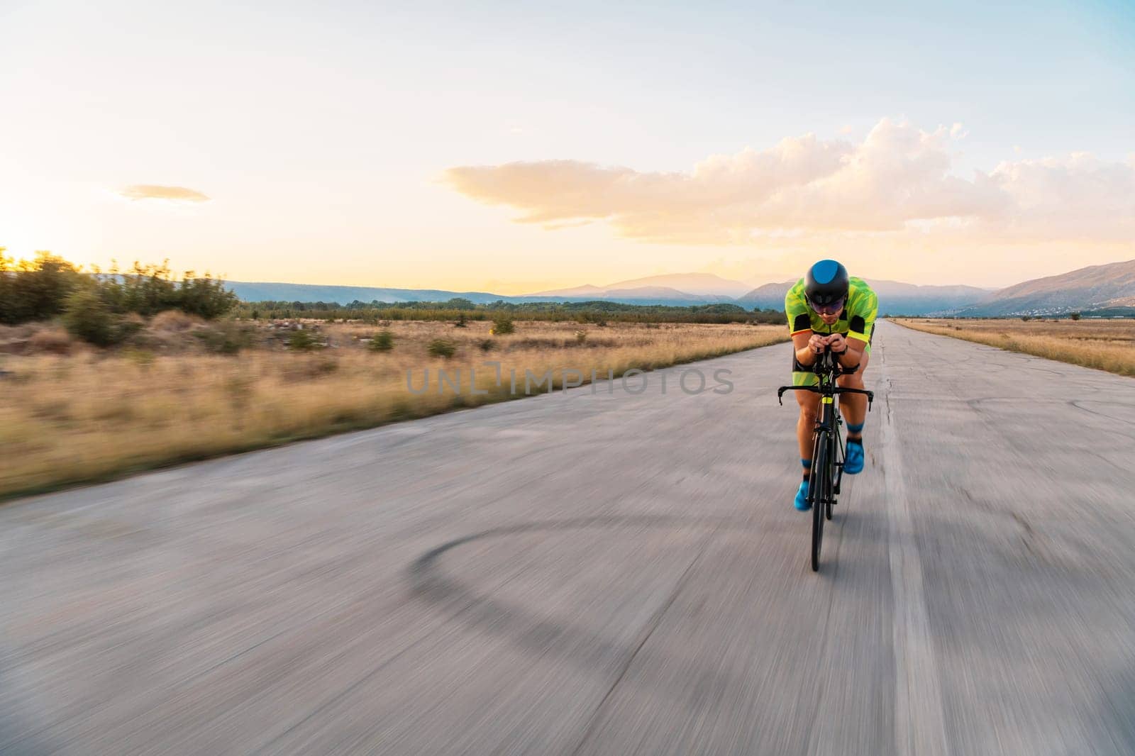 Triathlete riding his bicycle during sunset, preparing for a marathon. The warm colors of the sky provide a beautiful backdrop for his determined and focused effort