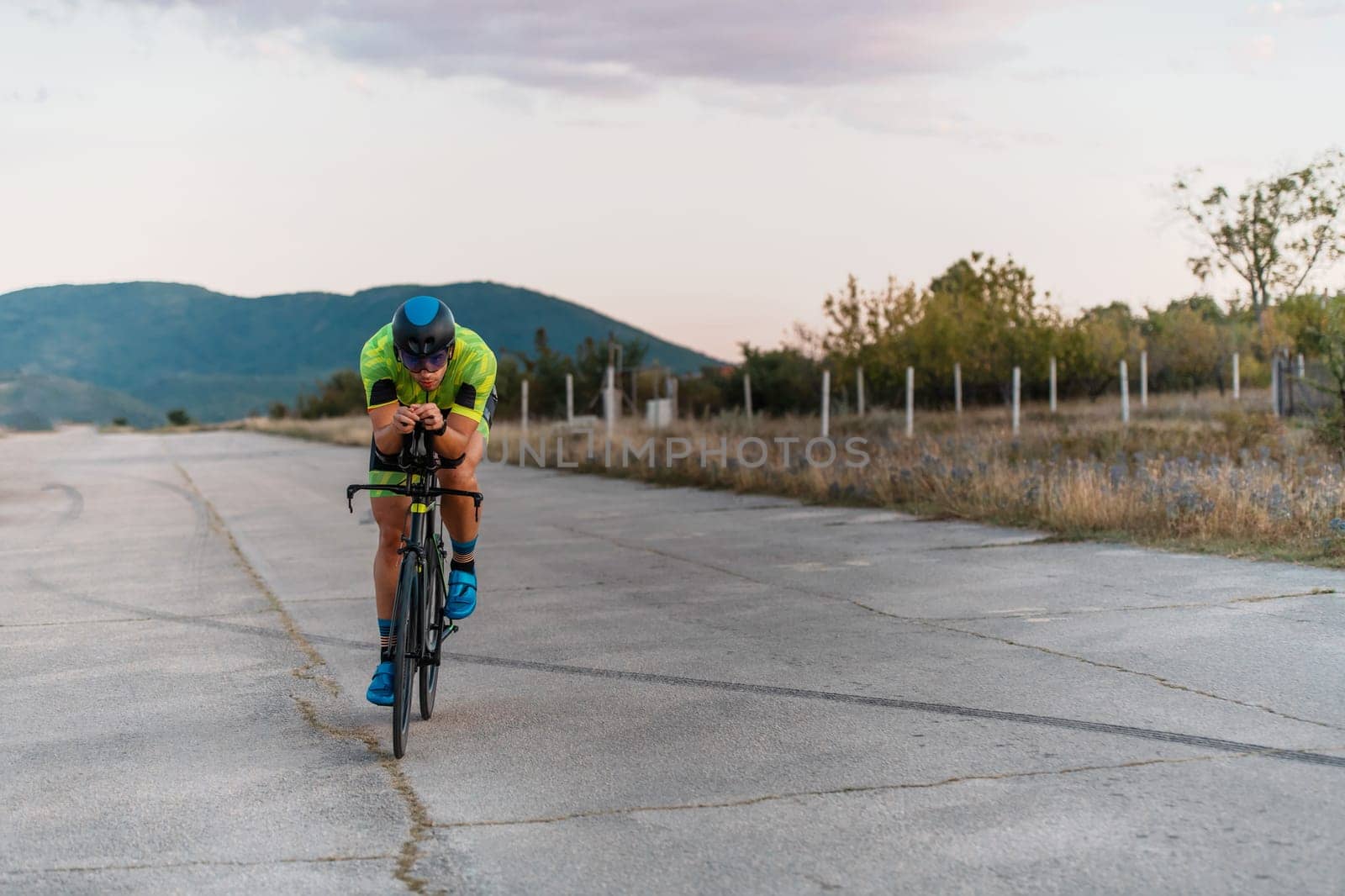Triathlete riding his bicycle during sunset, preparing for a marathon. The warm colors of the sky provide a beautiful backdrop for his determined and focused effort