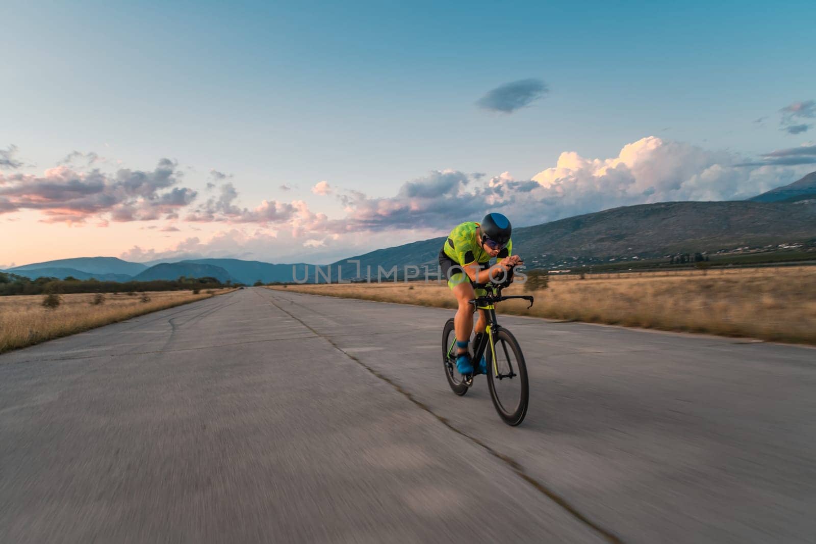 Triathlete riding his bicycle during sunset, preparing for a marathon. The warm colors of the sky provide a beautiful backdrop for his determined and focused effort