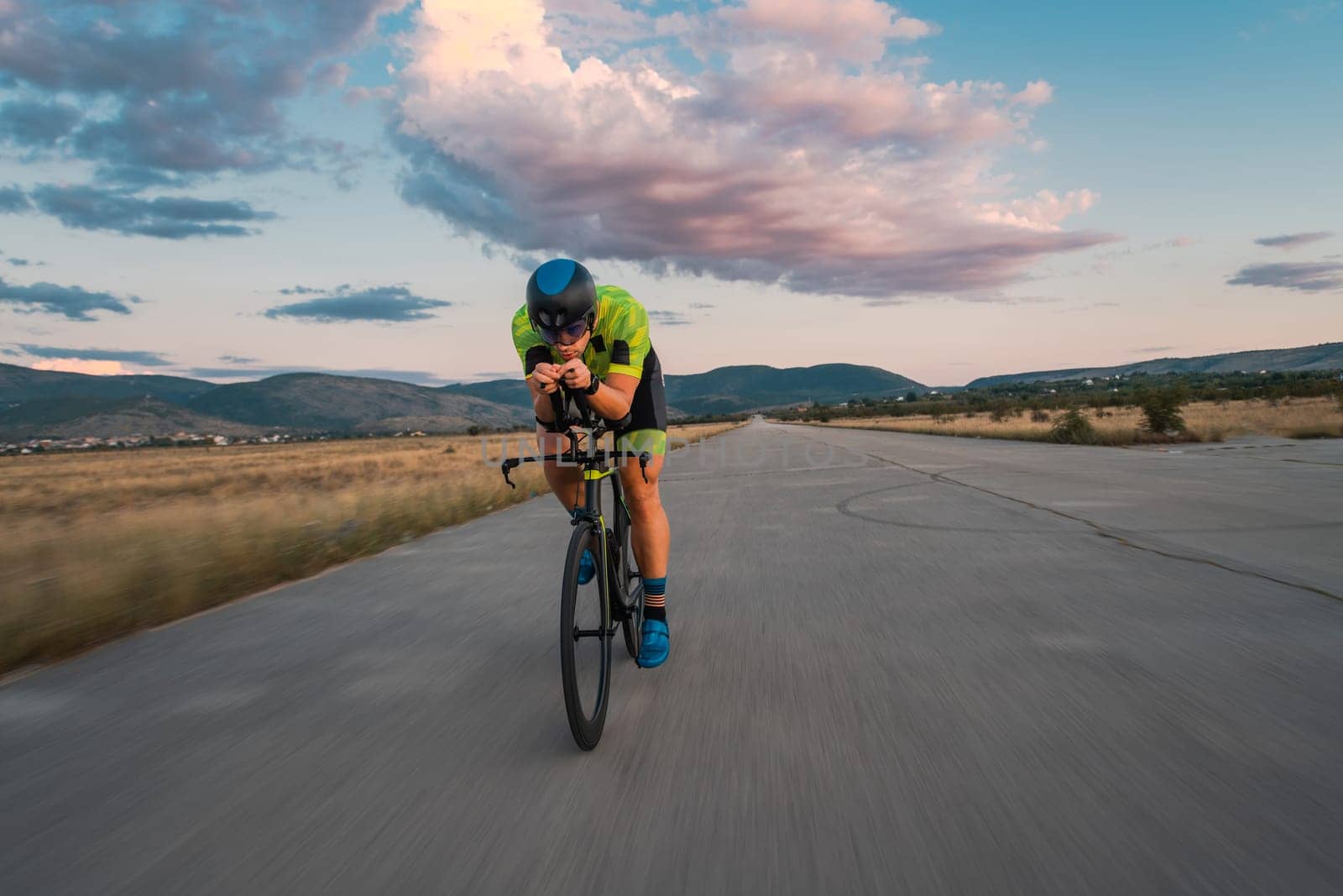Triathlete riding his bicycle during sunset, preparing for a marathon. The warm colors of the sky provide a beautiful backdrop for his determined and focused effort