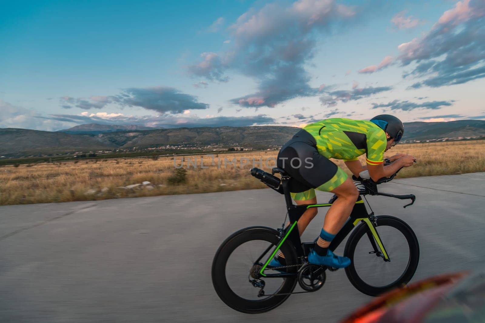 Triathlete riding his bicycle during sunset, preparing for a marathon. The warm colors of the sky provide a beautiful backdrop for his determined and focused effort