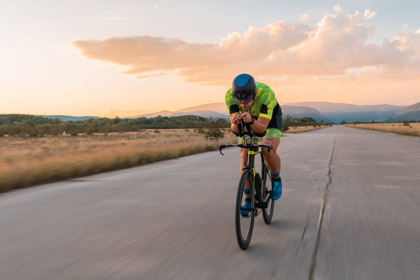 Triathlete riding his bicycle during sunset, preparing for a marathon. The warm colors of the sky provide a beautiful backdrop for his determined and focused effort