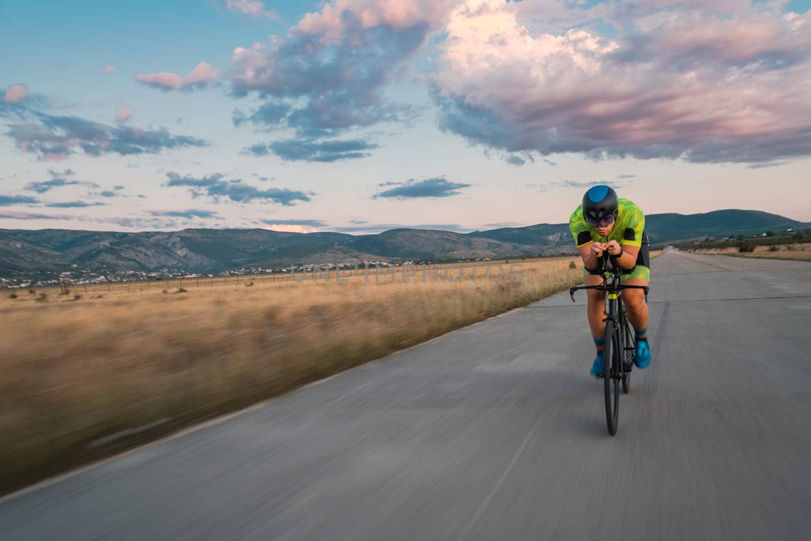 Triathlete riding his bicycle during sunset, preparing for a marathon. The warm colors of the sky provide a beautiful backdrop for his determined and focused effort