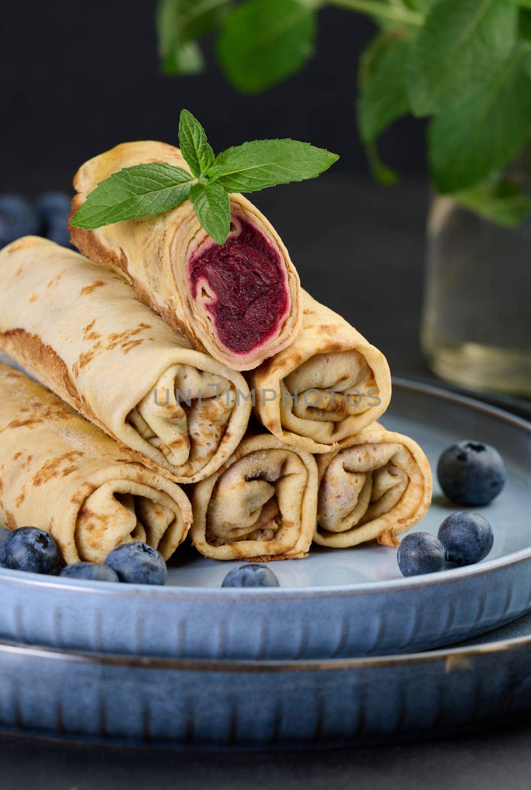 Crepes stacked in a pile, one of which is cut and filled with berry filling. The plate is also decorated with blueberries, black background