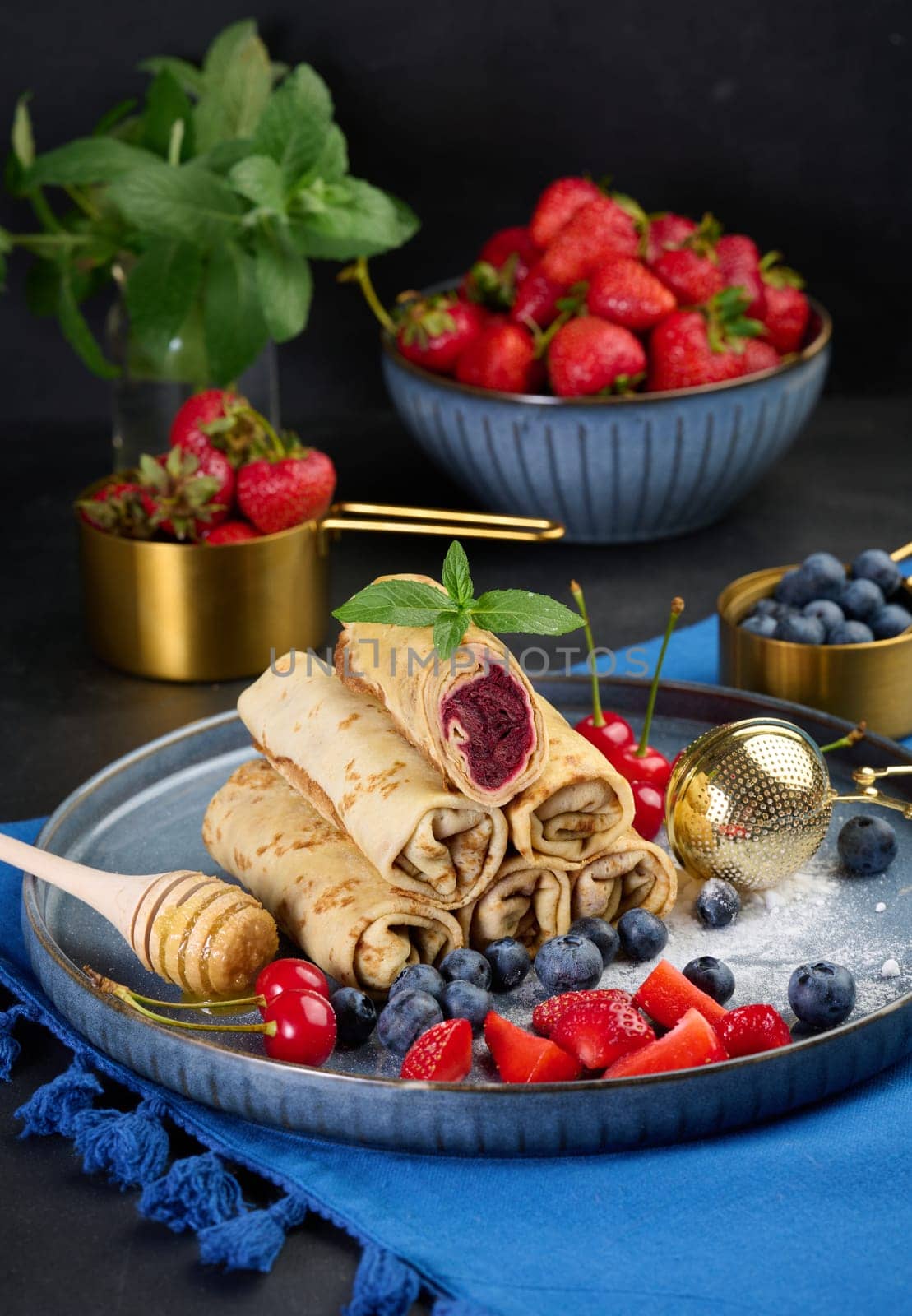 Crepes stacked in a pile, one of which is cut and filled with berry filling. The plate is also decorated with blueberries, strawberries, and cherries, a honey dipper with honey