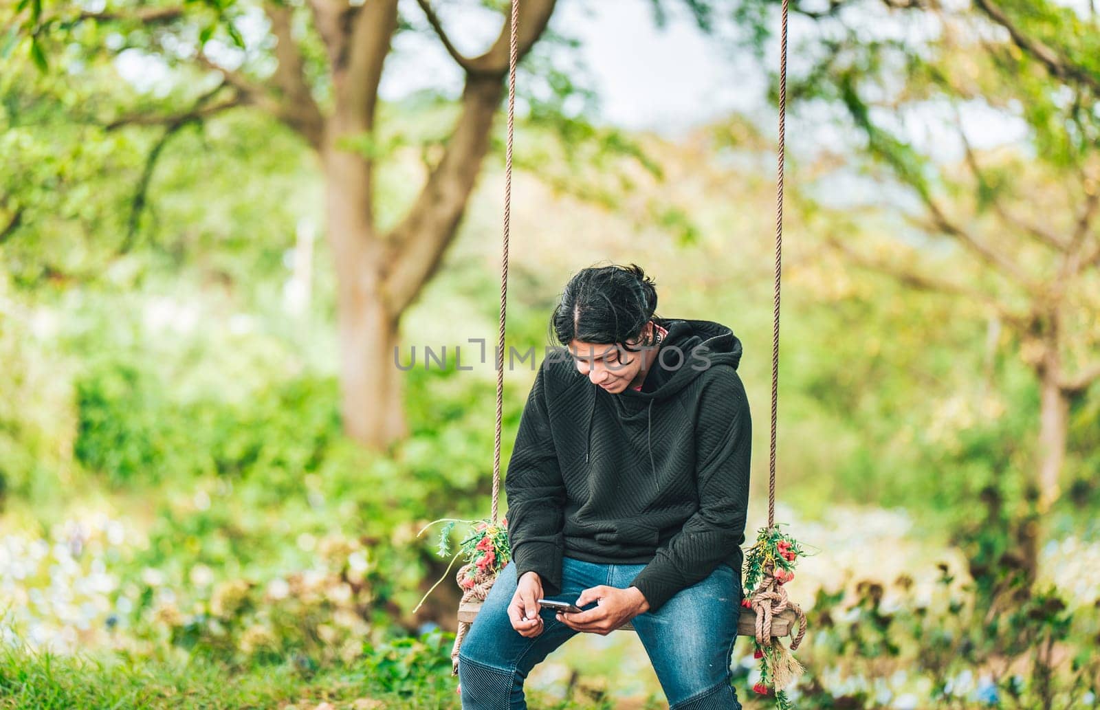 Smiling guy sitting on a swing with smart phone. Young man sitting on a swing using cell phone in nature