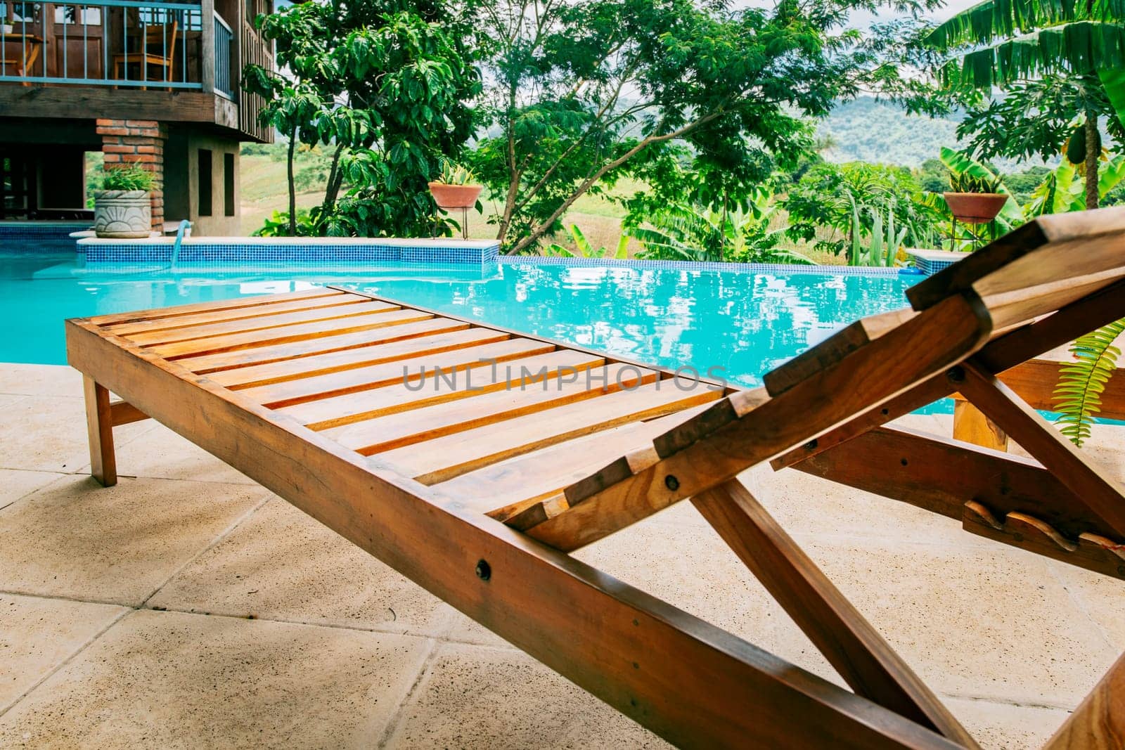 A wooden deck chair near a swimming pool. View of a Deck chair near a beautiful pool surrounded by nature