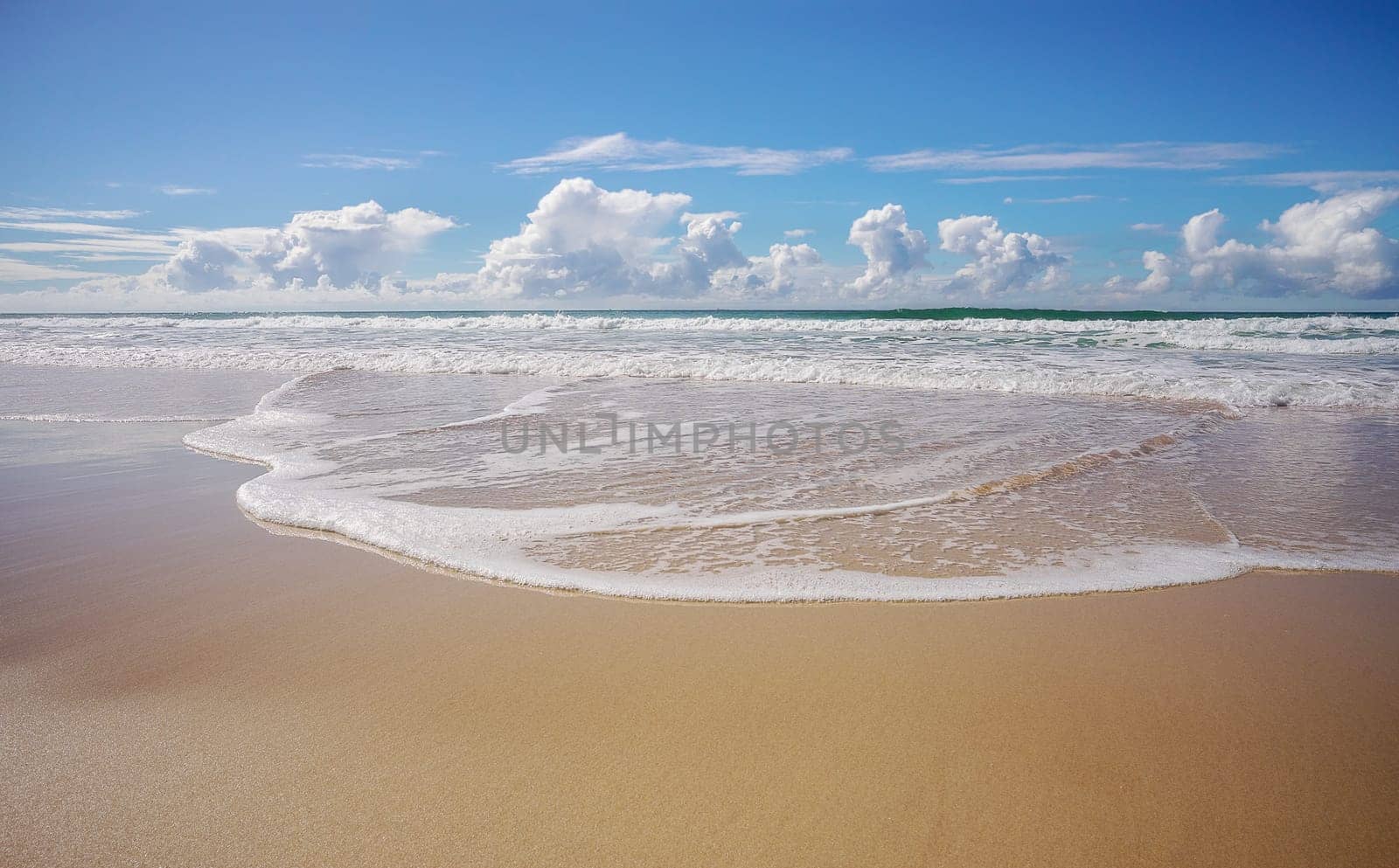 Tide coming in as waves break and roll onto seashore making foamy white patterns across the wet sandy beach, and  horizon with blue cloudy winter sky in background.
