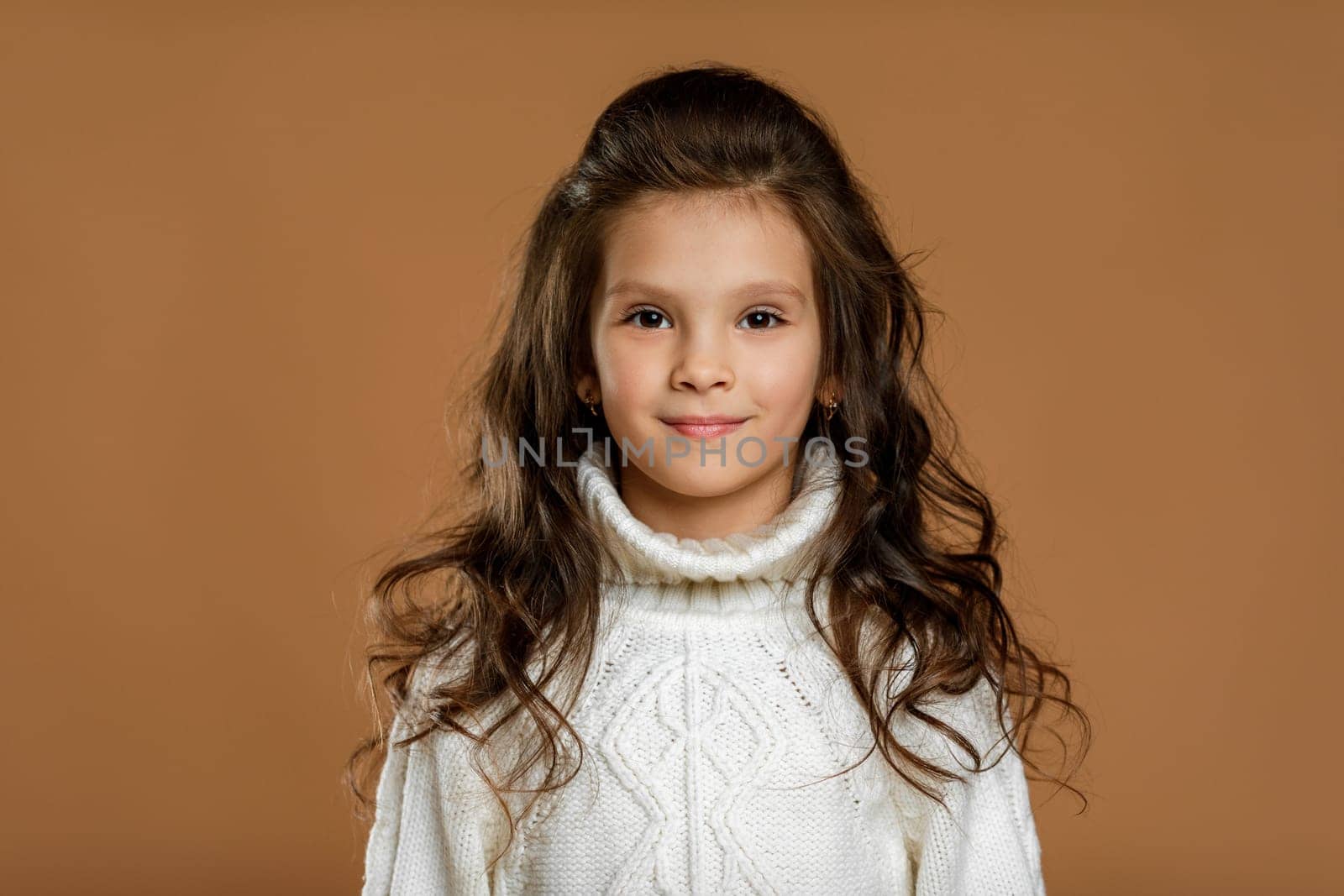 Cute smiling little child girl in white sweater looking to camera on beige background. Human emotions and facial expression