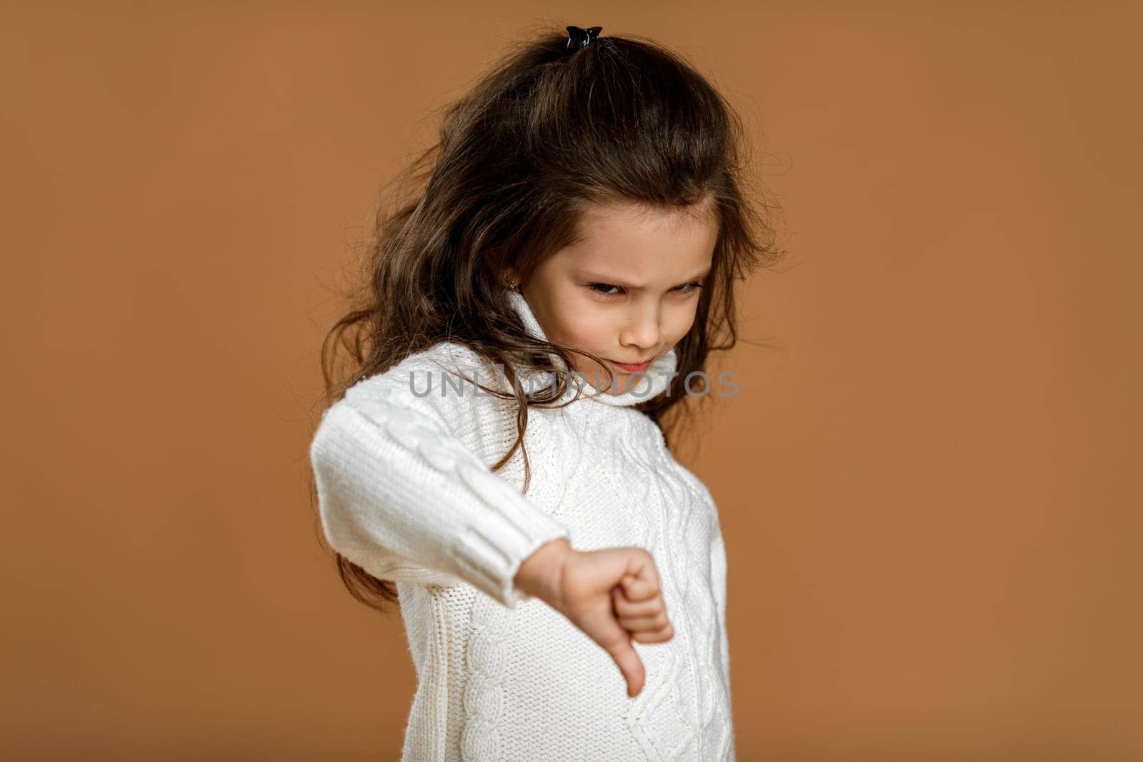 Cute little child girl in white sweater showing the thumbs down gesture on beige background.