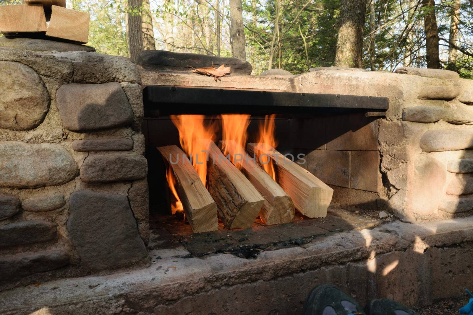 A stone outdoor fireplace with a fire burning, surrounded by trees.