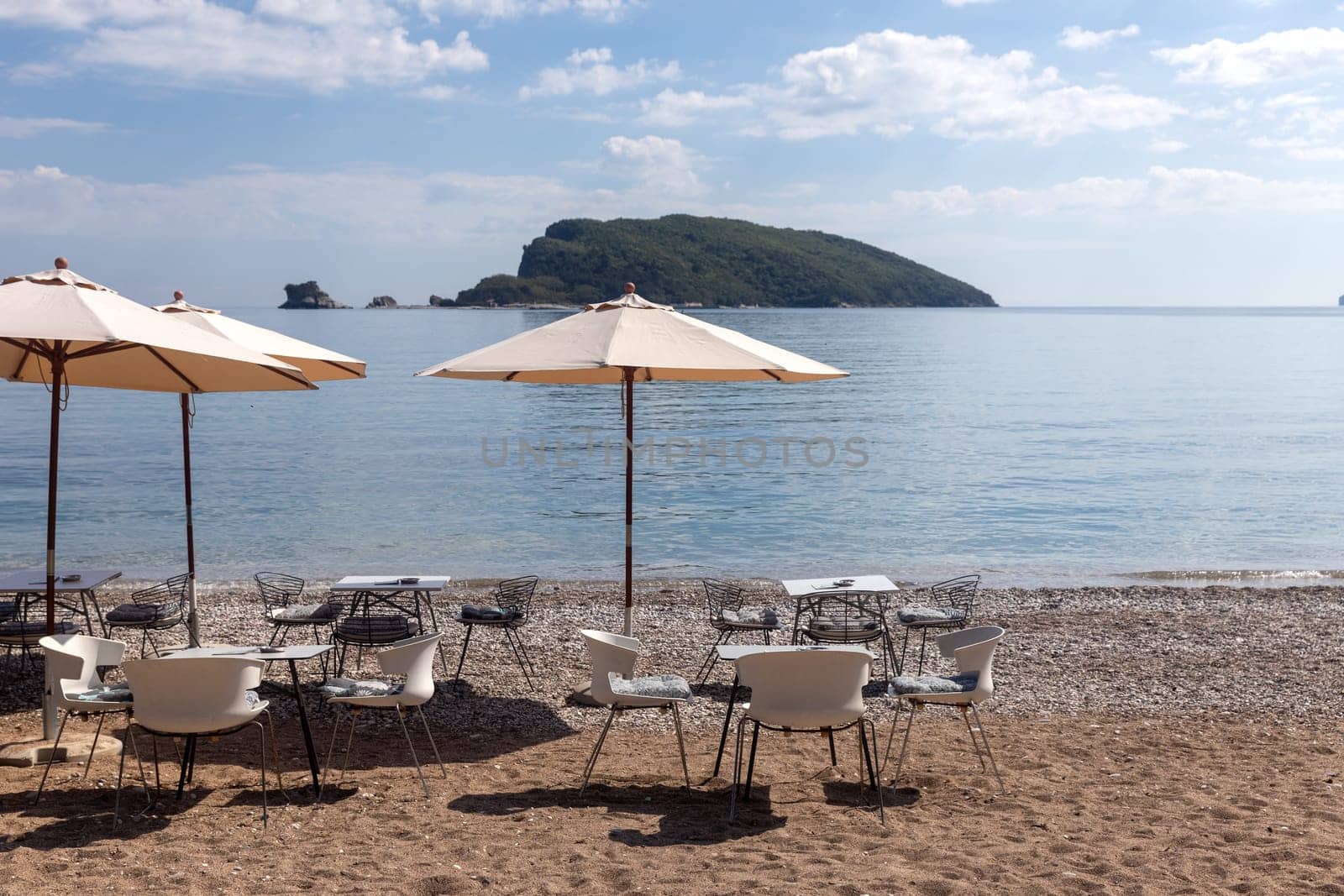 Open empty cafe on the beach with tables, chairs and umbrellas set up on the sand. Calm sea and island in the background.preparing for the season. Copy space.
