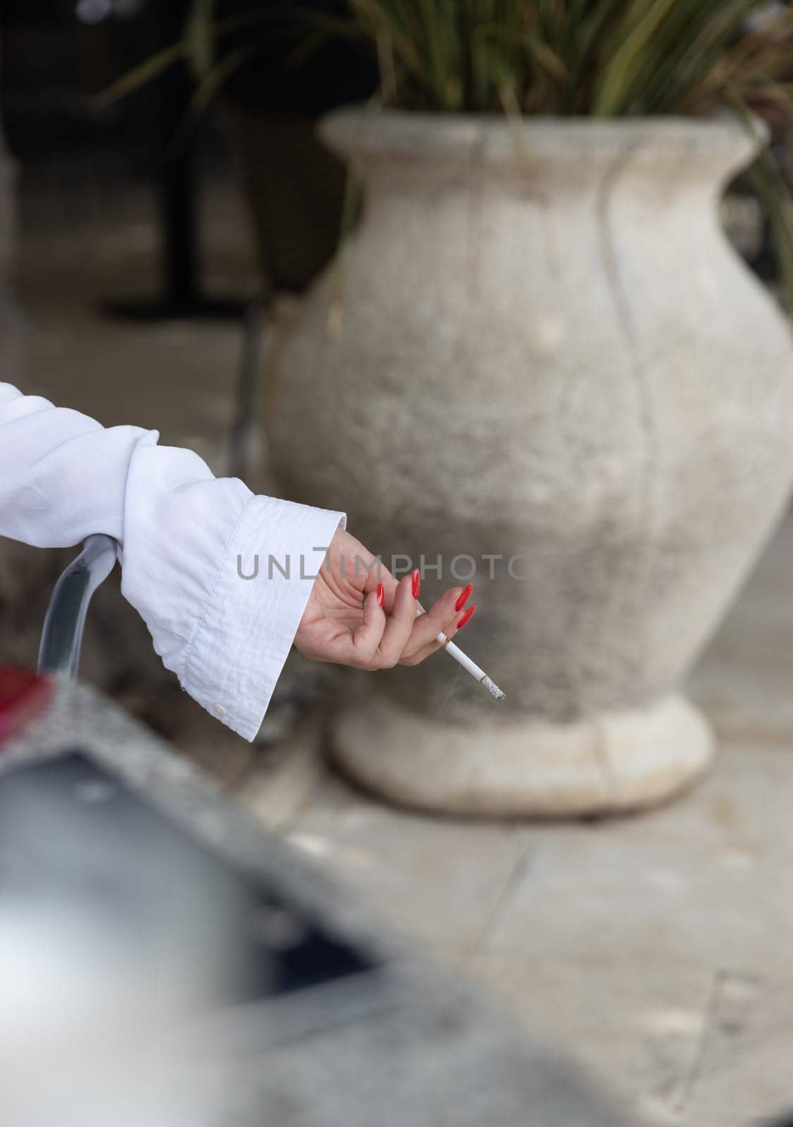 Female hand with red nails and white blouse holding a cigarette on blurred background of large ceramic pot outdoors.content related to lifestyle, habits and relaxation. High quality photo