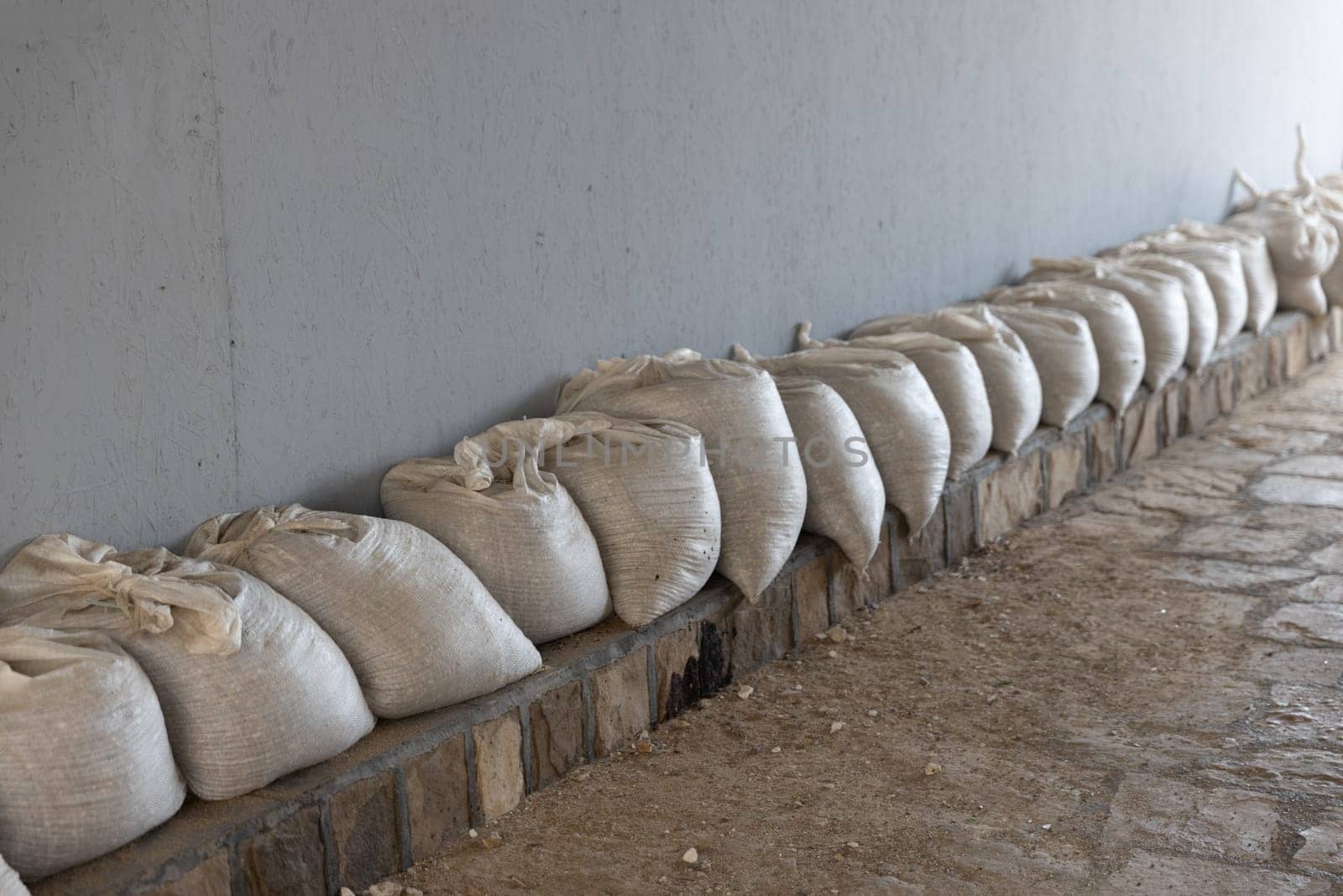 A row of white sandbags lined up against a wall on a stone floor for flood protection. The sandbags are neatly arranged against a plain wall. Copy space.