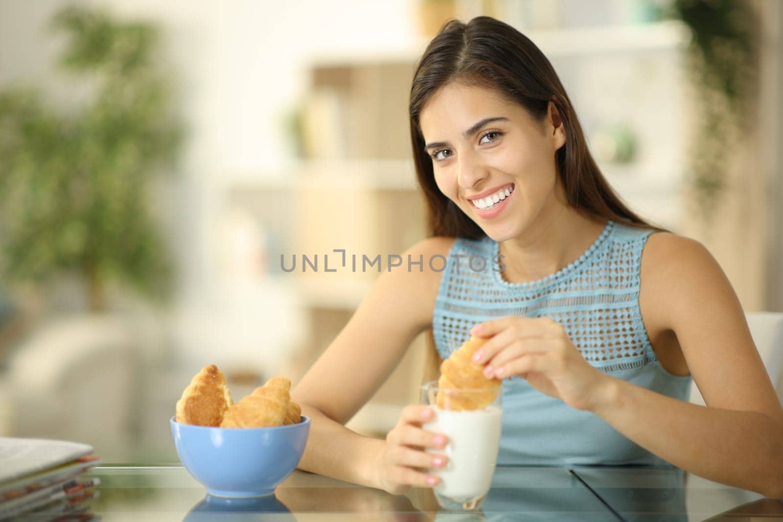 Happy woman dipping croissant in milk looks at camera at home