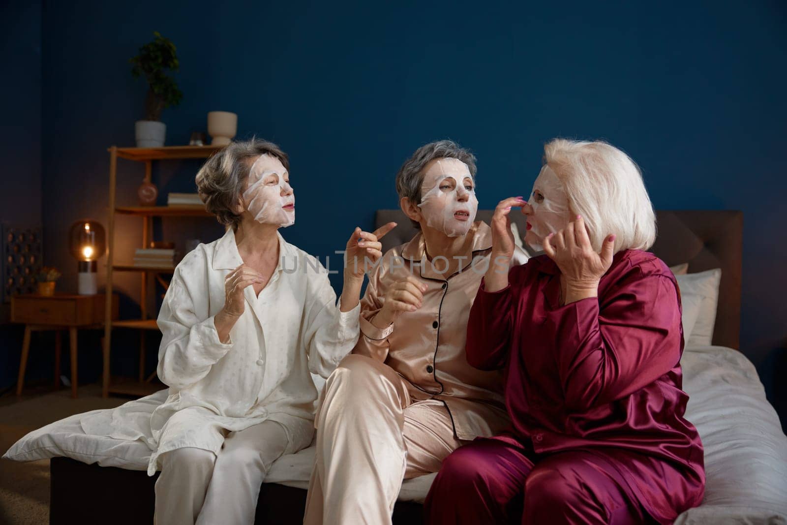 Senior women applying anti-aging cotton facial mask having fun together. Positive relaxed female best friends feeling cheerful and happy spending time on weekends