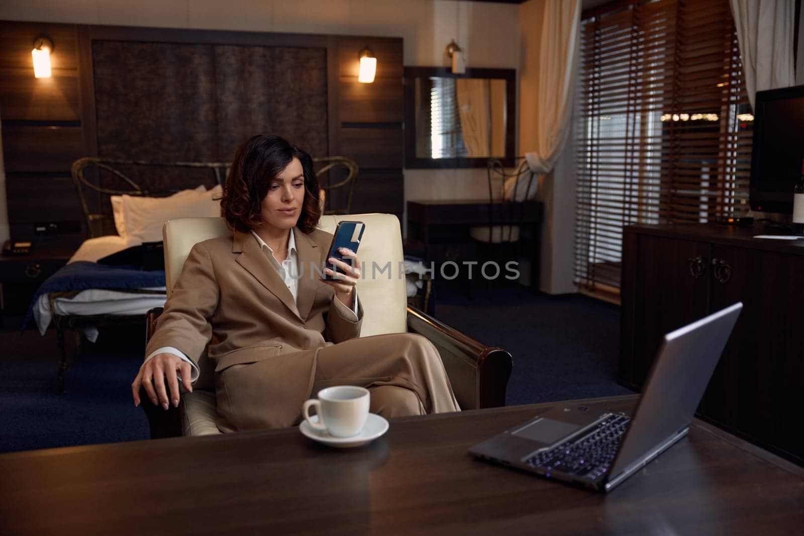 Young adult businesswoman using phone and laptop for work in luxury hotel room. Elegant female entrepreneur taking coffee break during busy job evening during business travel