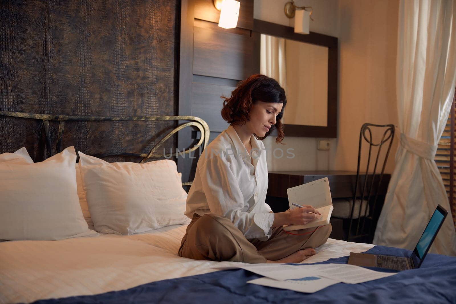 Busy young adult businesswoman working online from hotel room during business trip. Female entrepreneur doing research, preparing for financial report on laptop computer