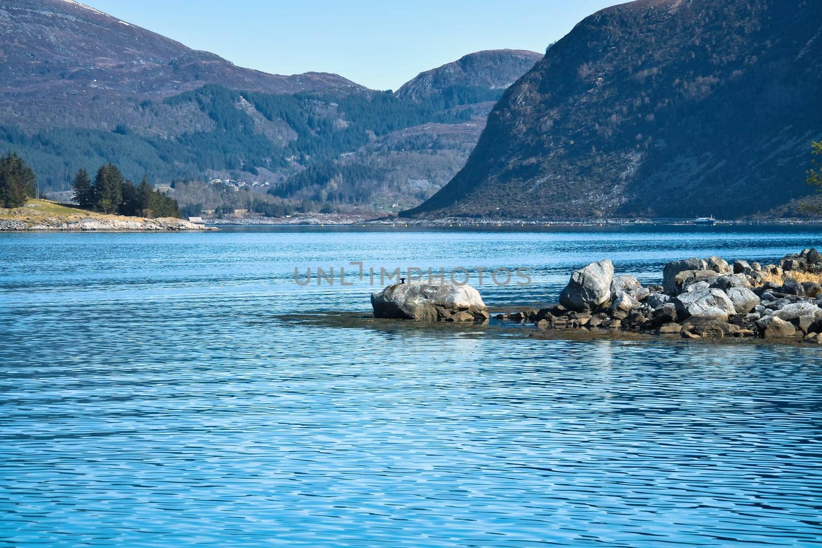 Fjord with mountains on the horizon. Stones reach into the water. The water glistens in the Norwegian sun. Landscape photo from the north