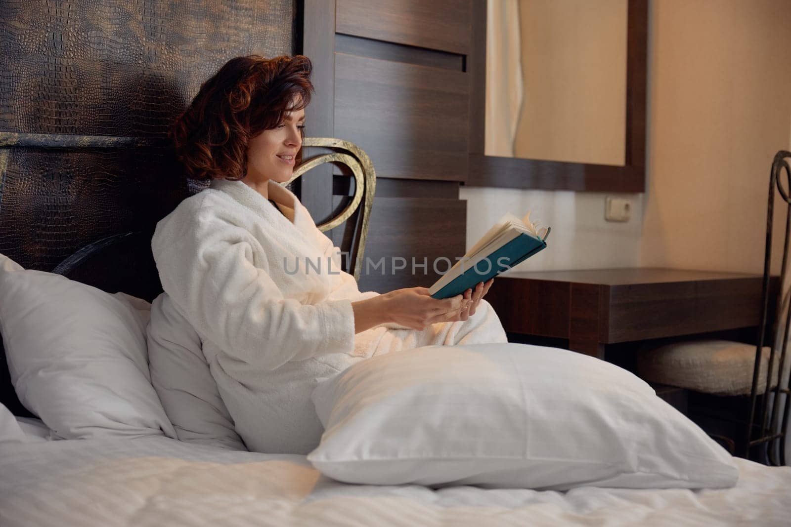 Businesswoman wearing bathrobe reading book lying in bed surrounded with soft pillow while rest in hotel room