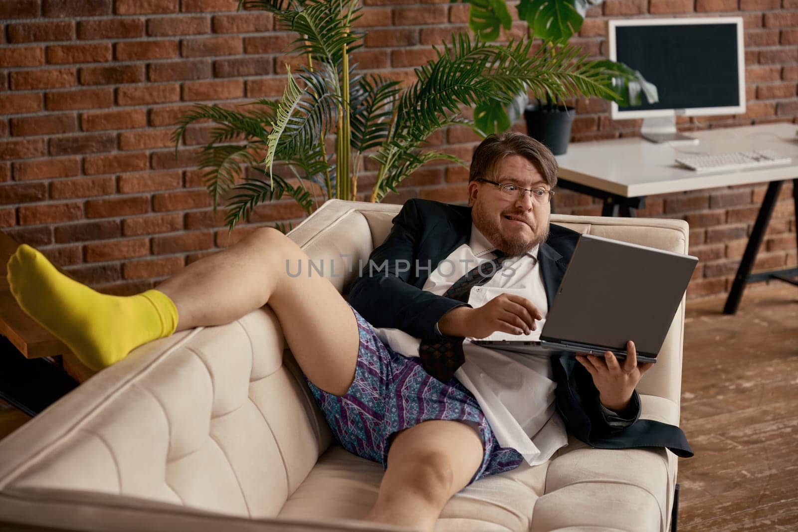 Surprised businessman grimacing looking at laptop screen working online sitting on sofa at home living-room. Confused salesman entrepreneur reading e-mail news using computer