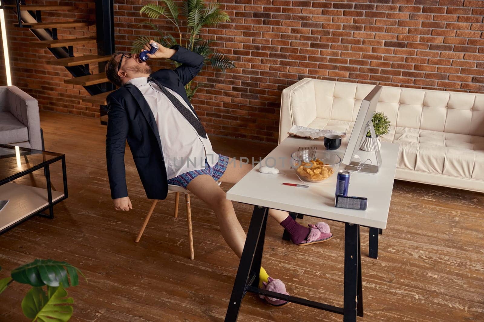 Funny businessman freelancer enjoying break time from online work at home office. Relaxed male entrepreneur eating snacks and drinking energy beverage while sitting at workplace