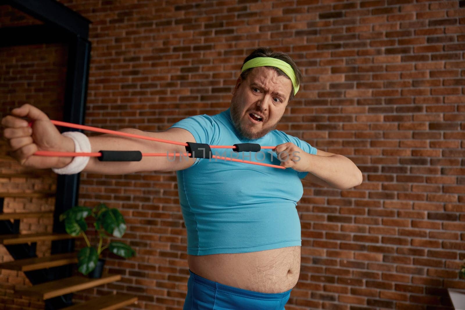 Comic chubby man in sportswear fooling around with fitness equipment during training at home. Fat sportsman crazy emotion