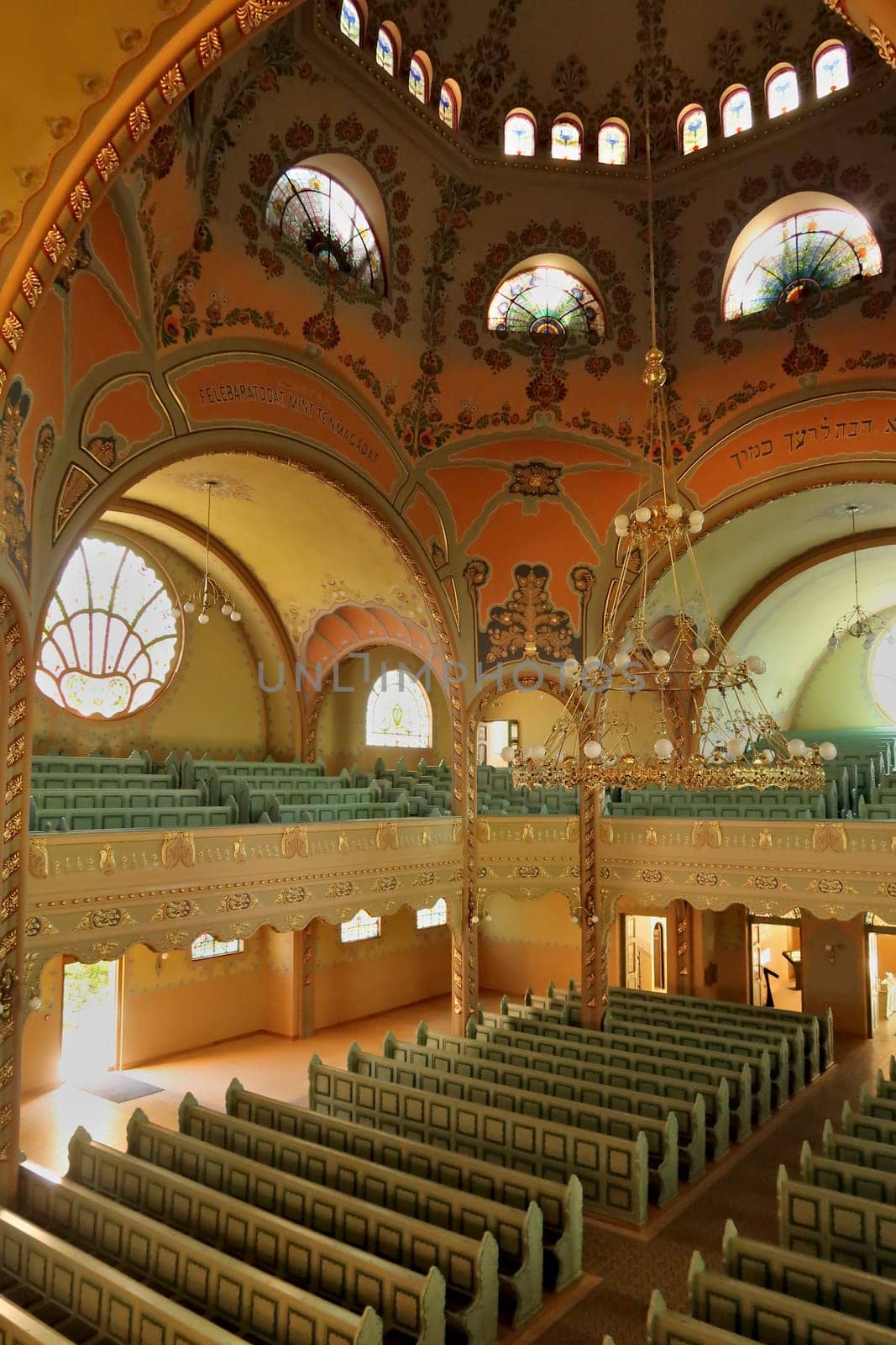 Inside the Synagogue of Subotica, a big golden chandelier is hanging from the painted ceiling over the benches and the aisle, light is shining through stained glass windows in the background, Subotica, Serbia 2022