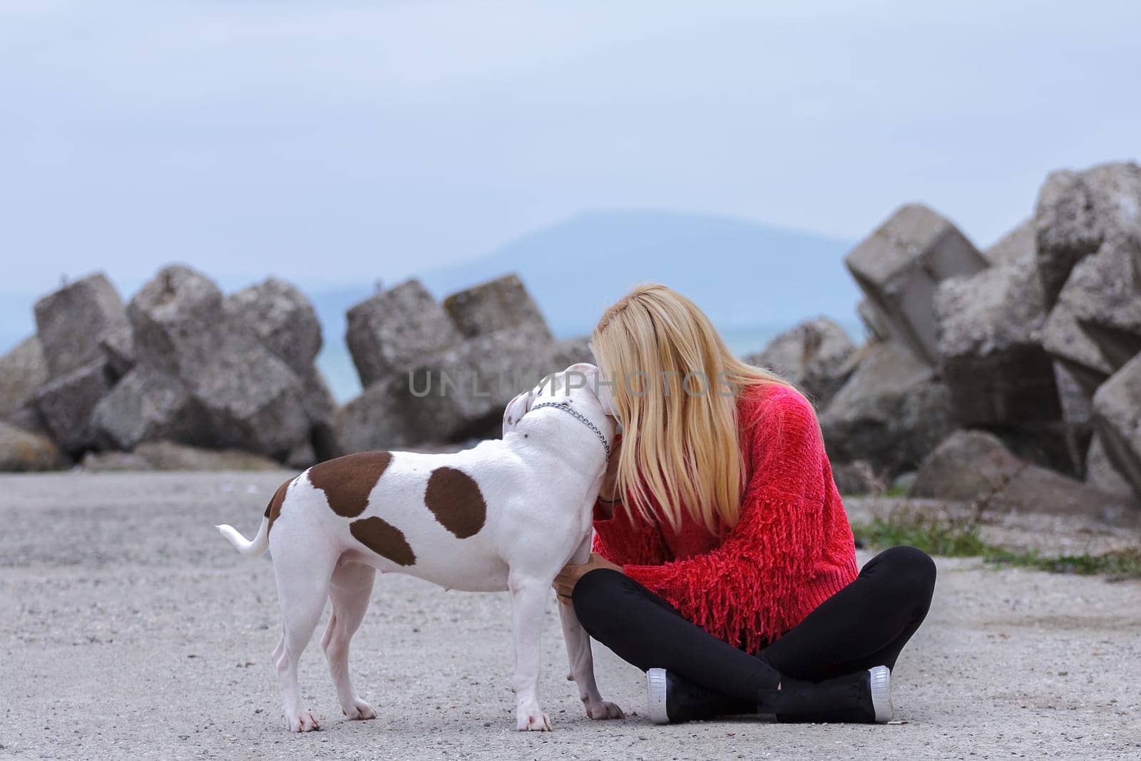 Sitting on the ground a blonde with a black wedge and a red blouse kisses a pit bull on the ear