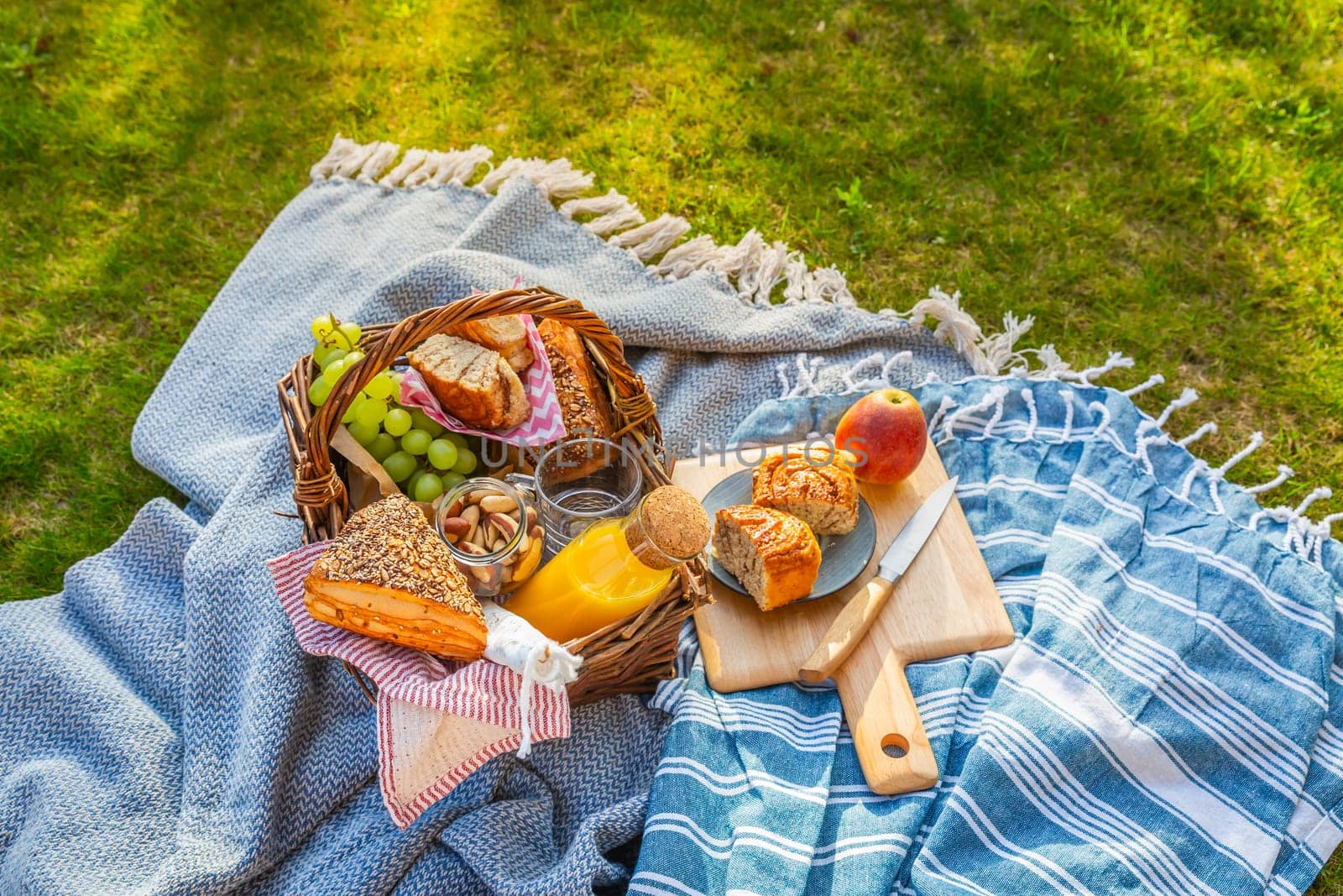 Picnic duvet and basket with different food, fruits, orange juice., yogurt and bread on green grass