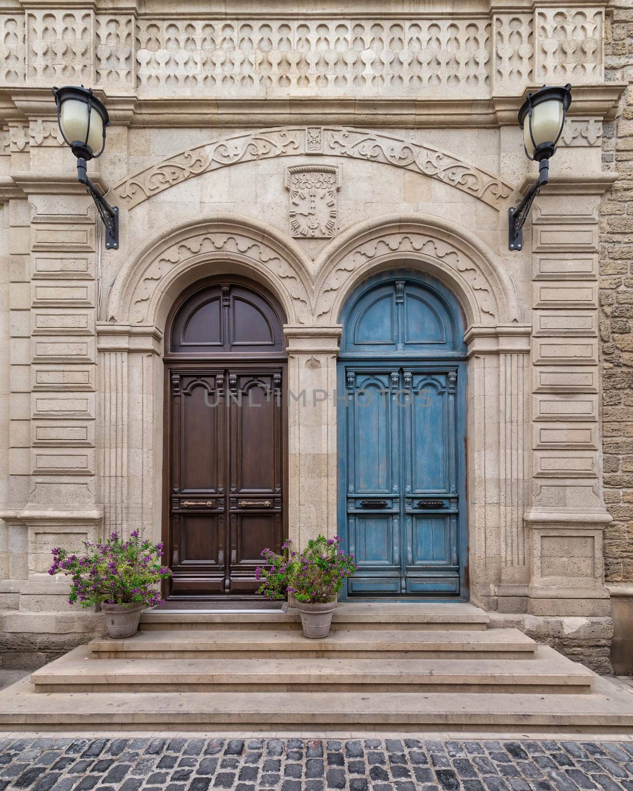 Showing a blue and a brown wooden doors side by side, this picture highlights the contrast between two different architectural styles. The building is decorated with street lights and plants
