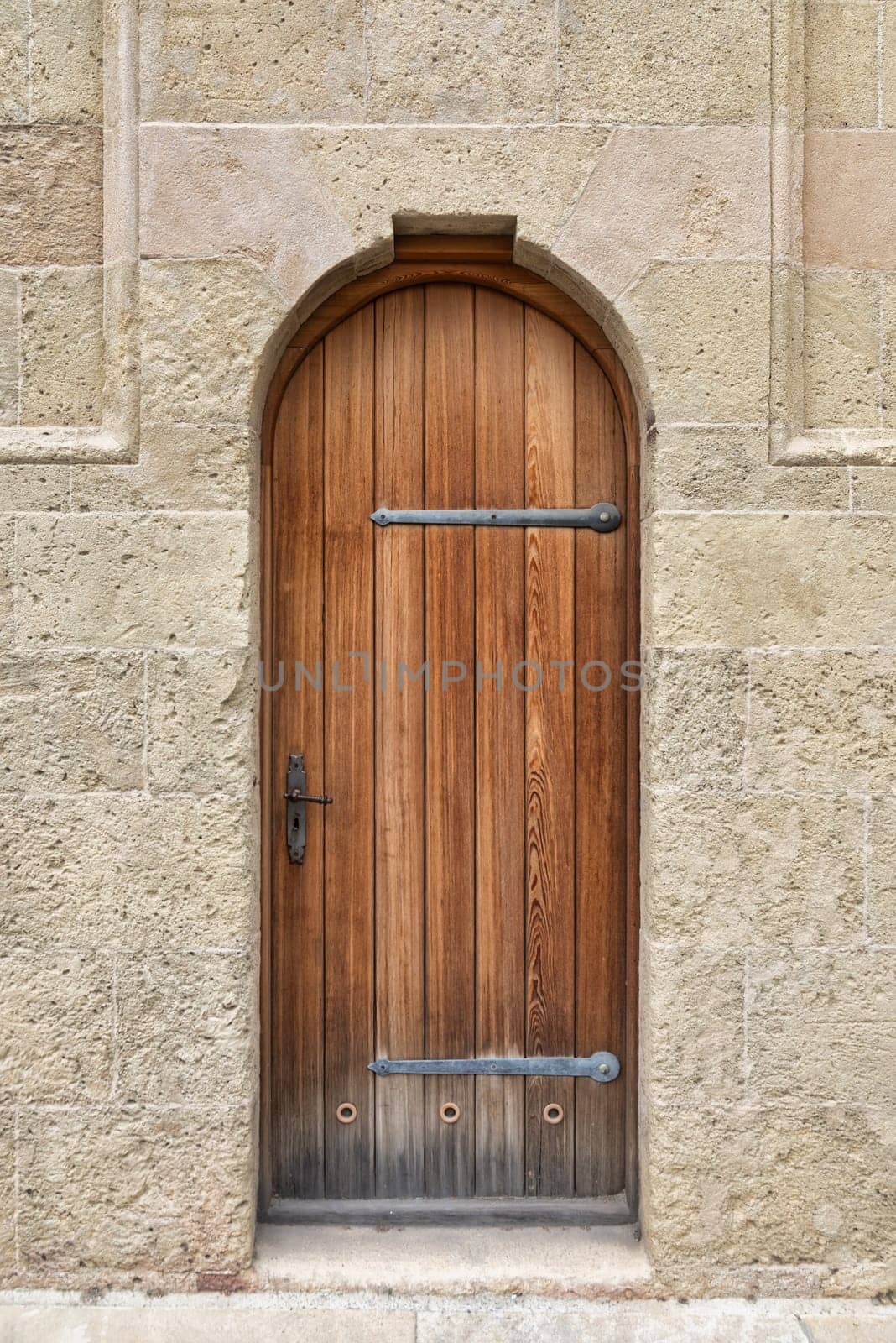 A close-up image of a weathered, arched wooden door set within a stone wall. The door is made of dark brown wood with horizontal planks and has a metal handle and two large metal hinges