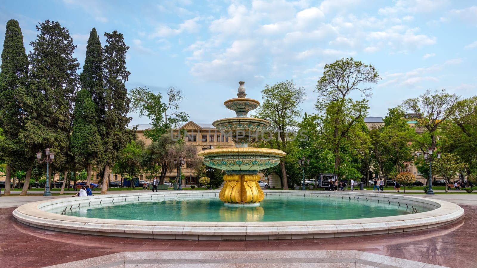 Baku, Azerbaijan - May 5, 2024: In Bakus Sahil Bagi park, a golden based fountain with blue and white tiles sits in a circular pool, surrounded by lush trees and a walkway