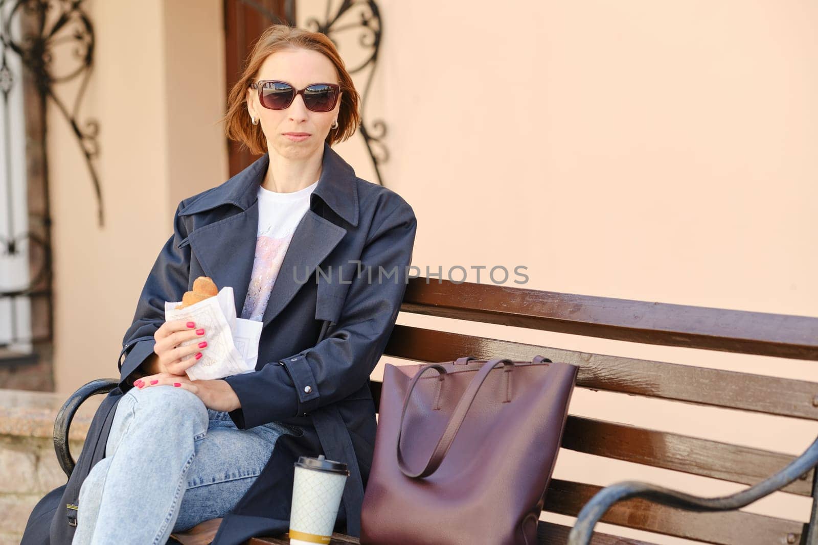 Redhead woman enjoys cappuccino and croissant on city bench in stylish outfit.