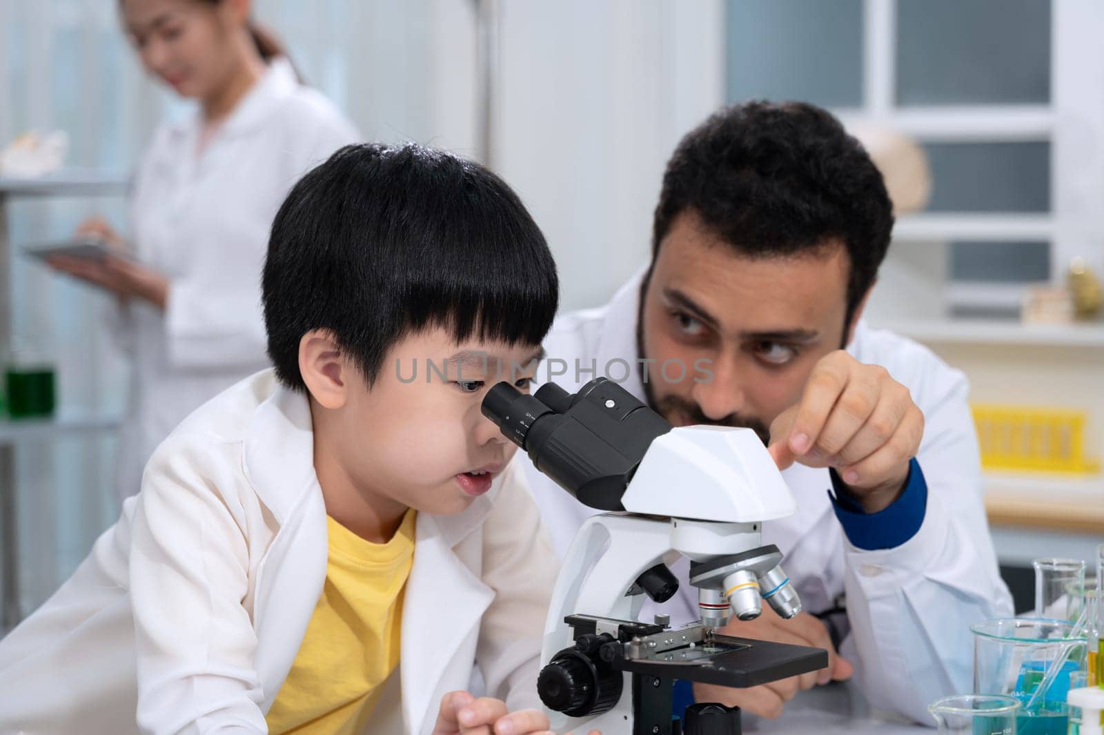 Kid boy wearing white laboratory coat and learning how to use microscope in science laboratory room with scientist teacher. Children education concept with experiment, fun and enjoy class.