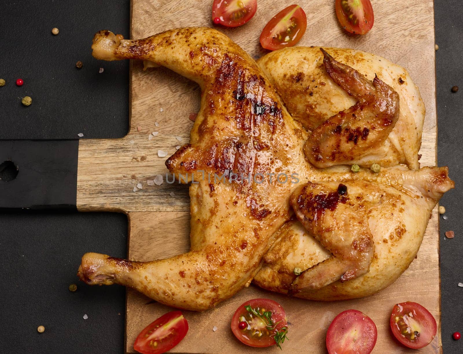 Whole fried chicken with spices on a wooden board on a black table, top view