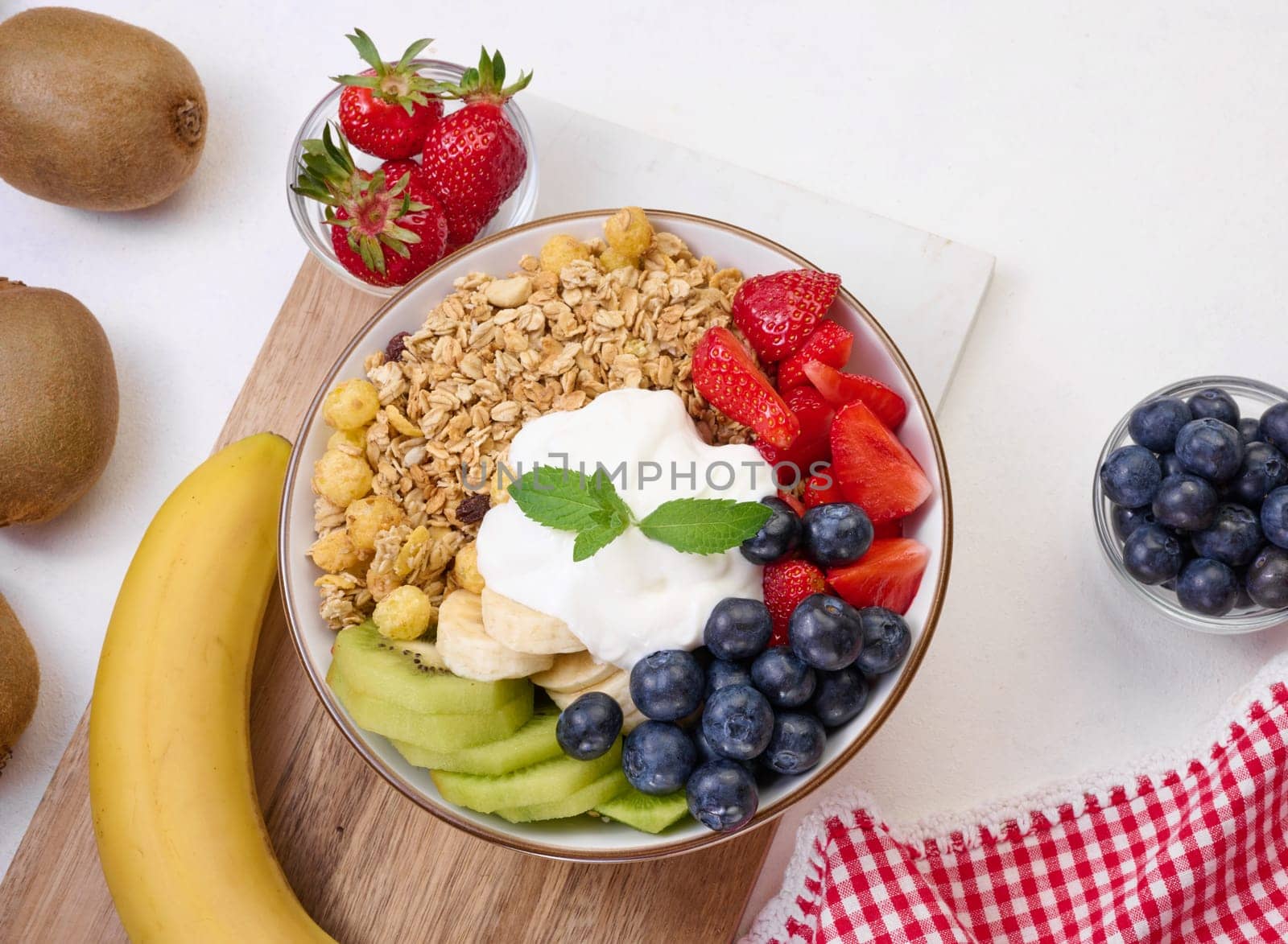 Granola with strawberries, kiwi, banana and blueberries in a round plate on a white table. Healthy and tasty food, top view