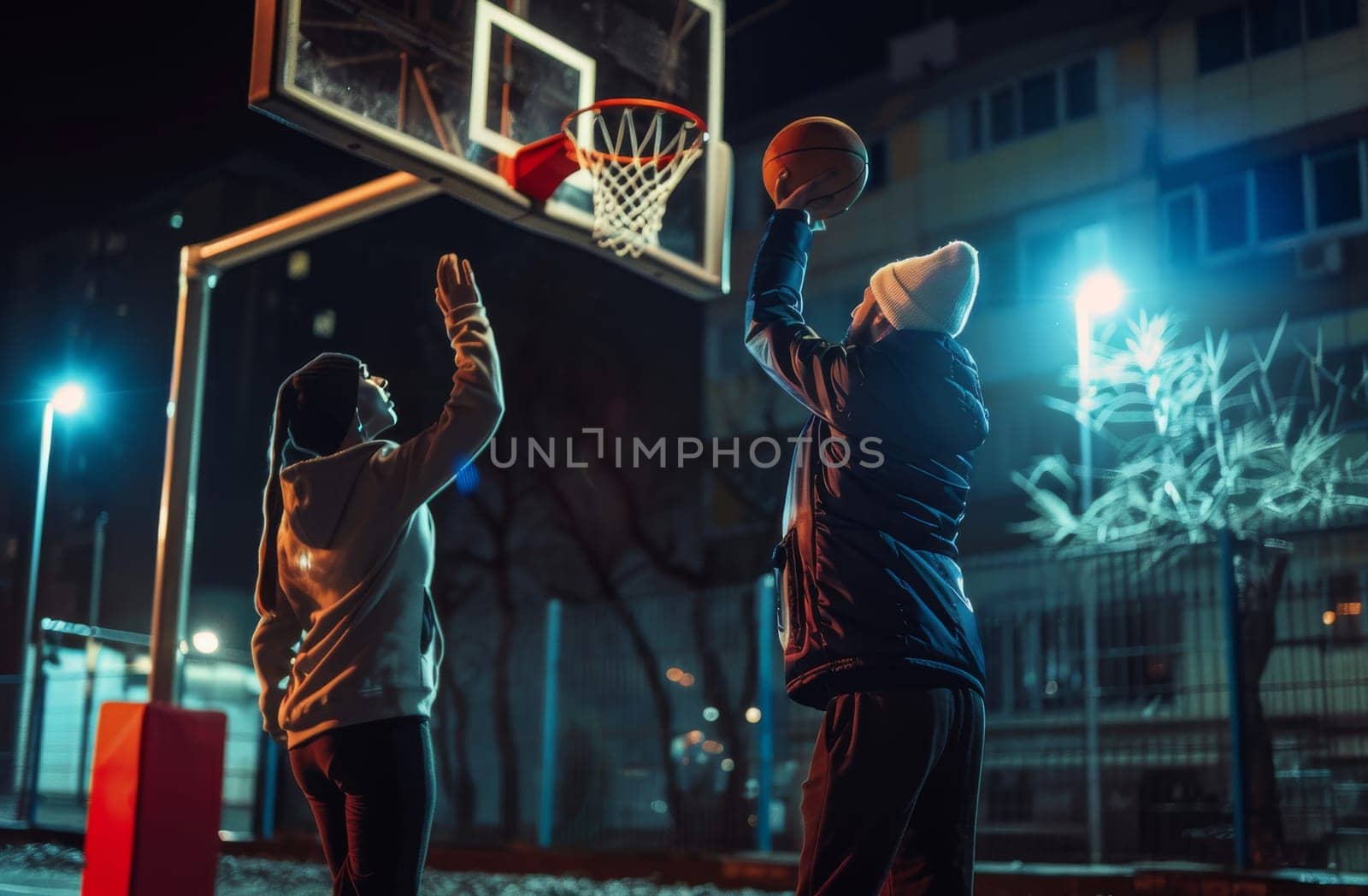 Two people are playing basketball at night under street lights, with one shooting the ball towards the hoop