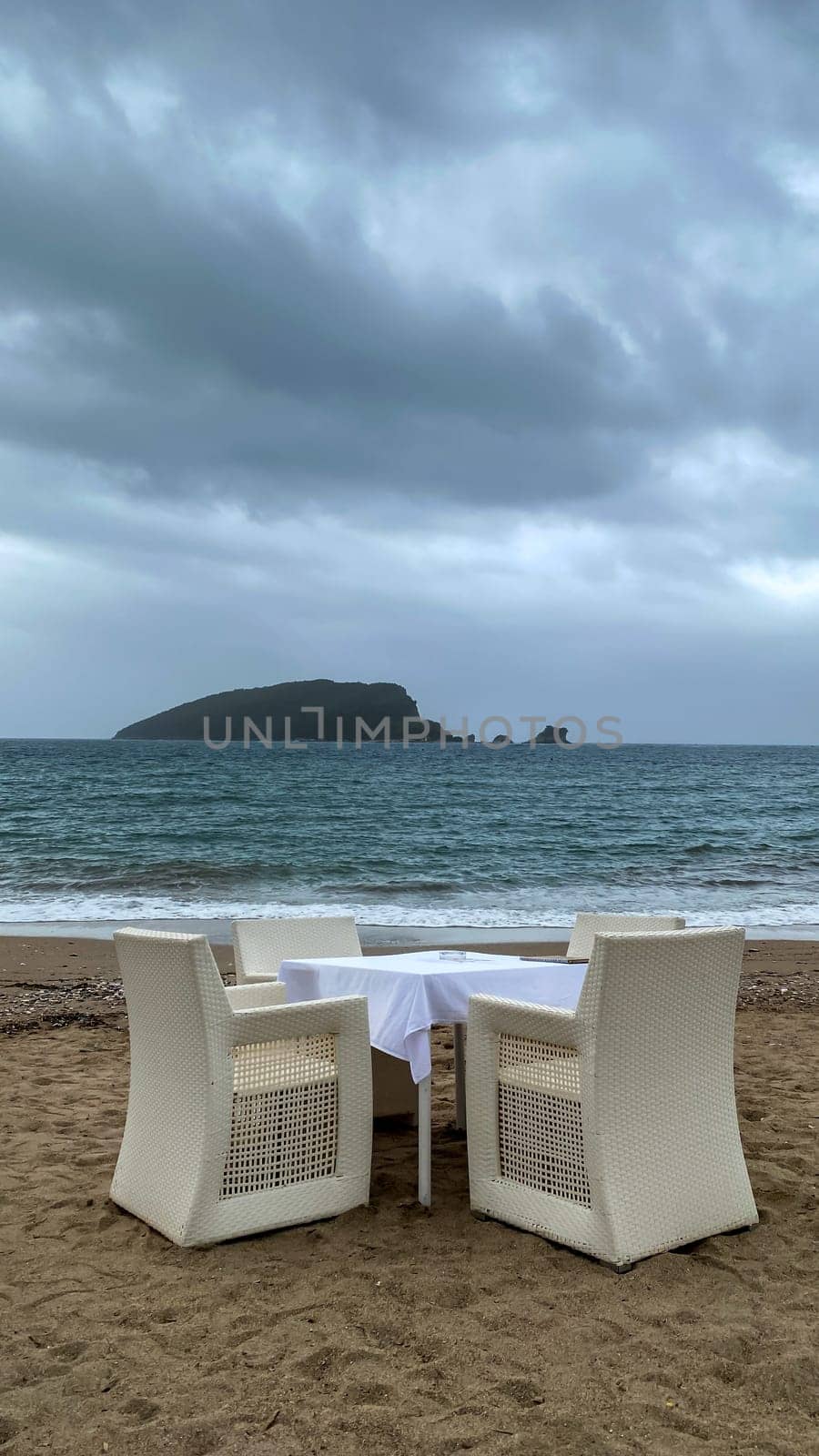 A table with a blue checkered tablecloth and white chairs set on a sandy beach, overlooking the turquoise sea and a distant island. Overcast sky adds a serene atmosphere. Copy space.