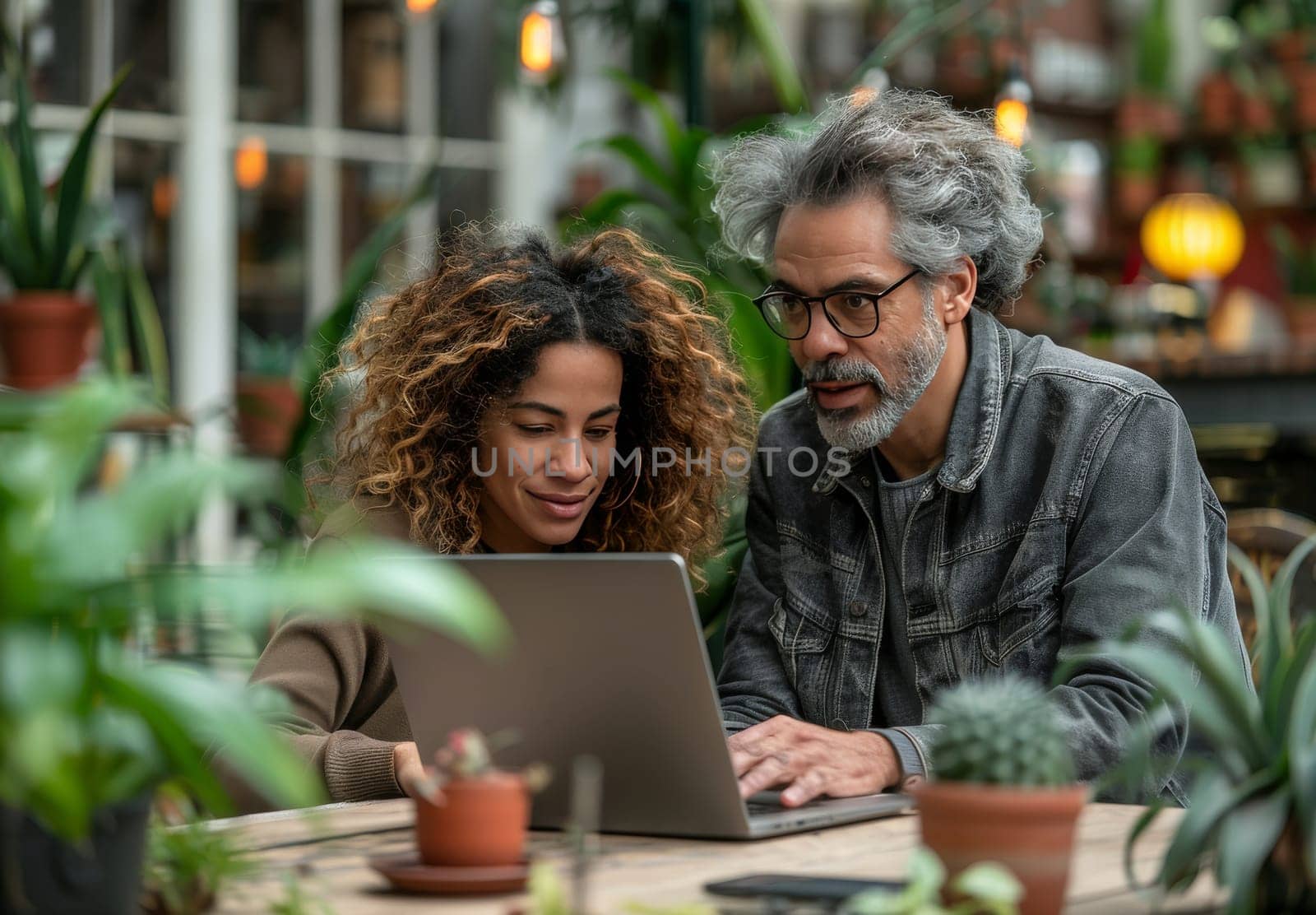 A man and woman in their thirties, both wearing glasses, are seated at a table in a lush greenhouse, their focus intently on a laptop screen. Sunlight streams through the glass roof, casting a warm glow over the couple, suggesting an intimate and shared moment.