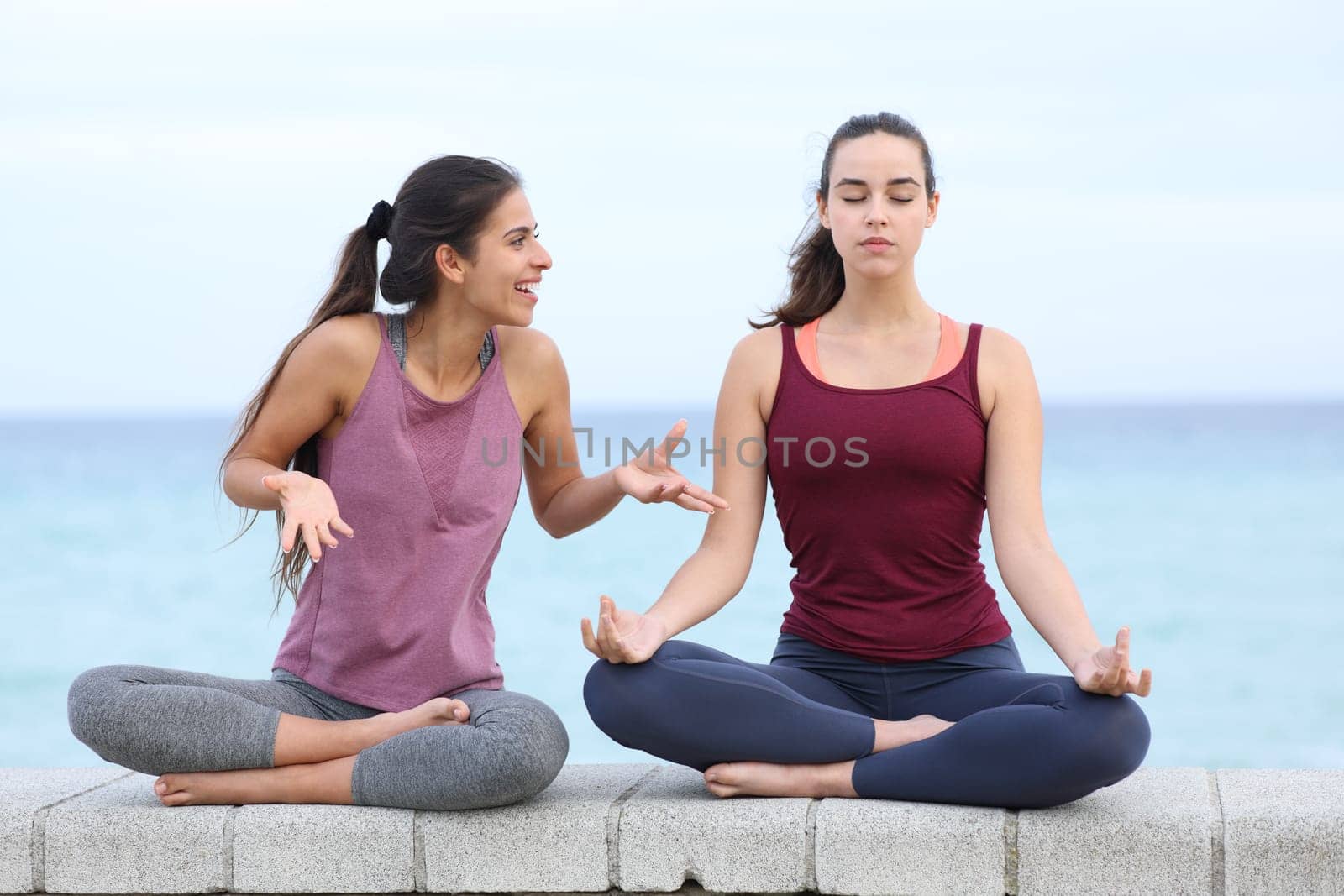 Front view portrait of a yogi trying to concentrate doing yoga and a friend disturbing on the beach