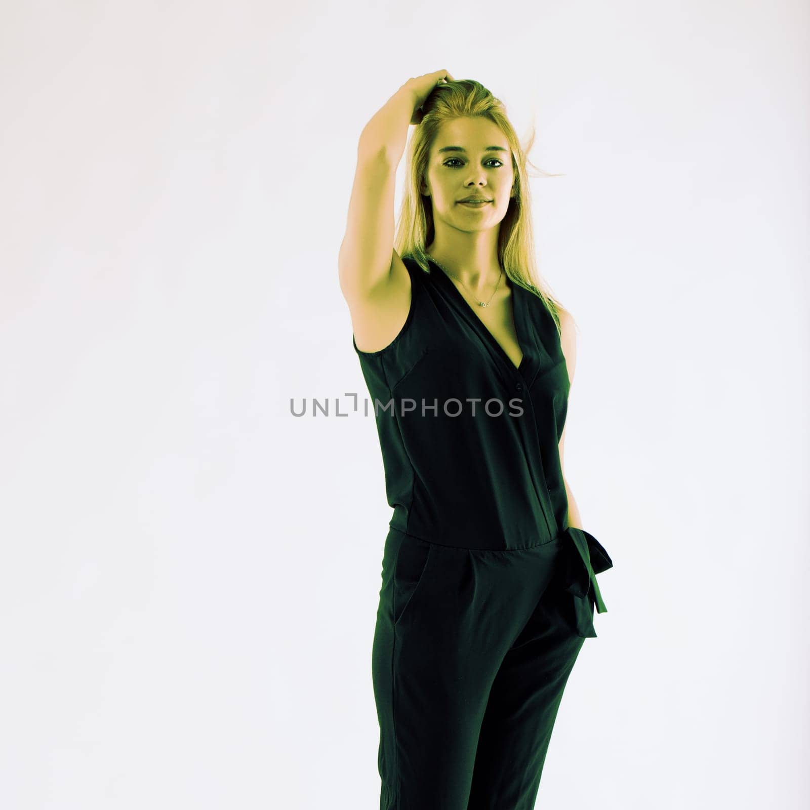 Attractive young woman with long blonde hair is posing in a studio, wearing a black overall, touching her hair with her hand